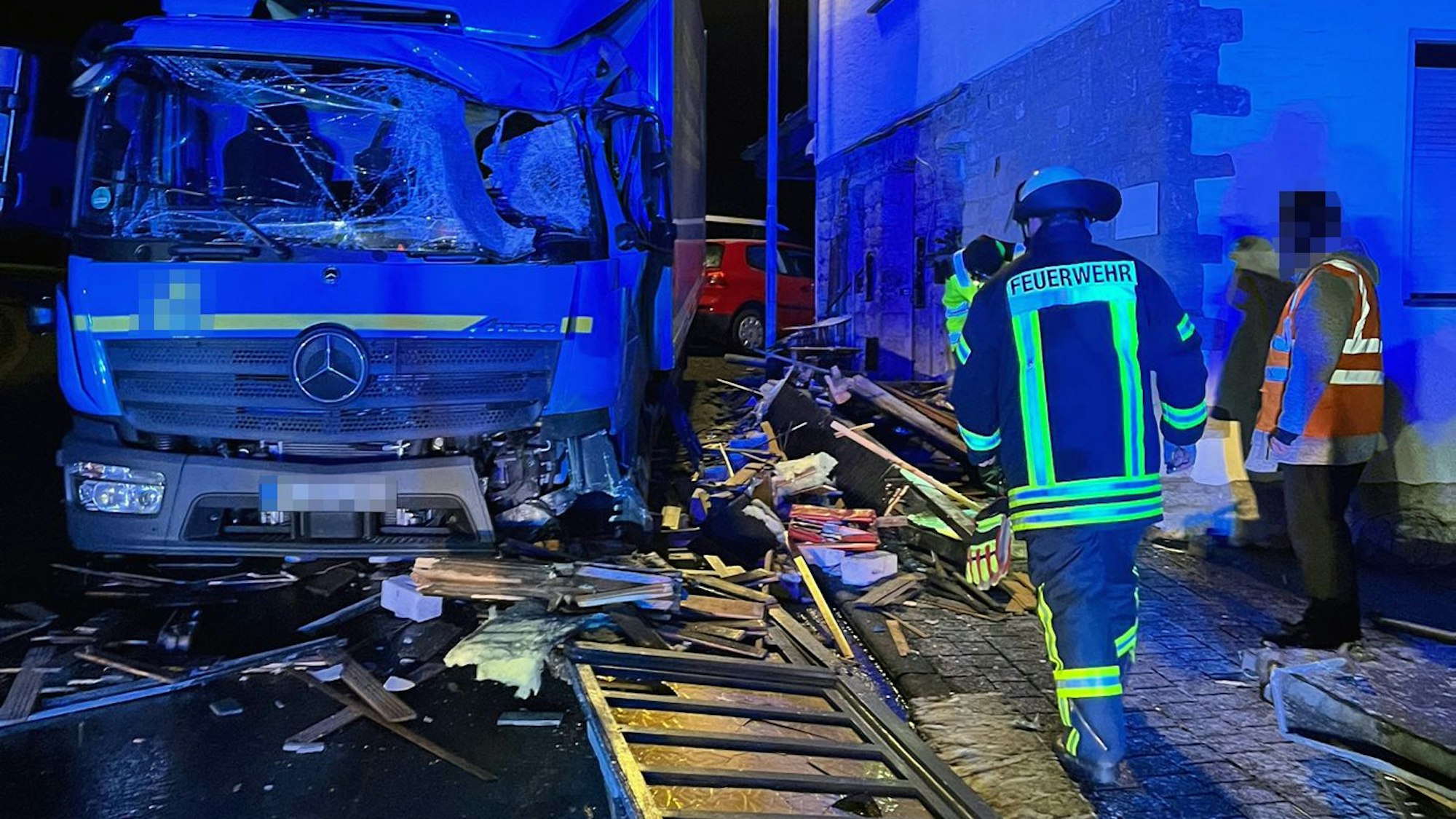 Ein Lkw steht mit stark beschädigter Windschutzscheibe auf einer Straße, vor ihm liegen Holzlatten. Feuerwehrleute sind vor Ort.