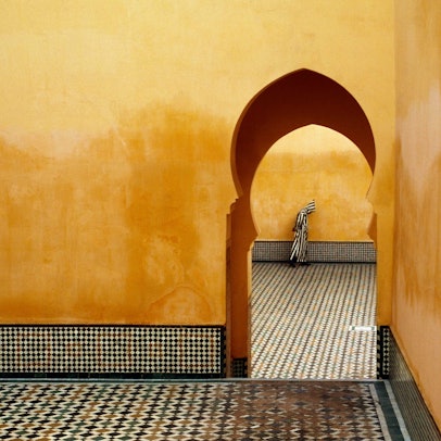 Die Aufnahme „MOROCCO. Meknes. Moulay Ismael Mausoleum (Muslim shrine). 1985“ zeigt den Blick durch ein Tor, im Hintergrund steht ein Mann mit einer gestreiften Kutte mit Kapuze.