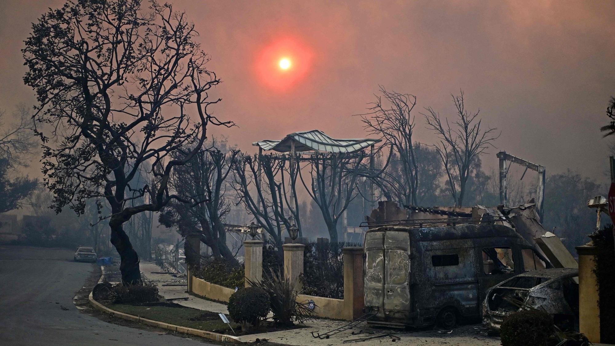 TOPSHOT - The sun is seen behind smoke above charred structures and vehicles after the passage of the Palisades Fire in Pacific Palisades, California, on January 8, 2025. At least five people have been killed in wildfires rampaging around Los Angeles, officials said on January 8, with firefighters overwhelmed by the speed and ferocity of multiple blazes. (Photo by AGUSTIN PAULLIER / AFP)