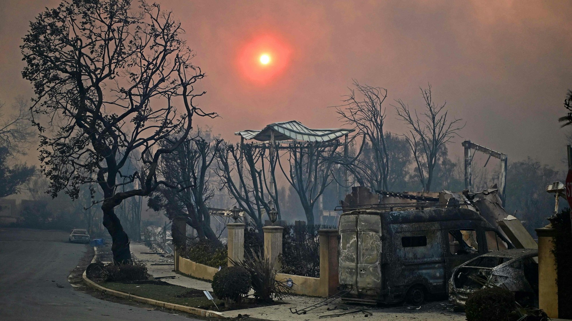TOPSHOT - The sun is seen behind smoke above charred structures and vehicles after the passage of the Palisades Fire in Pacific Palisades, California, on January 8, 2025. At least five people have been killed in wildfires rampaging around Los Angeles, officials said on January 8, with firefighters overwhelmed by the speed and ferocity of multiple blazes. (Photo by AGUSTIN PAULLIER / AFP)