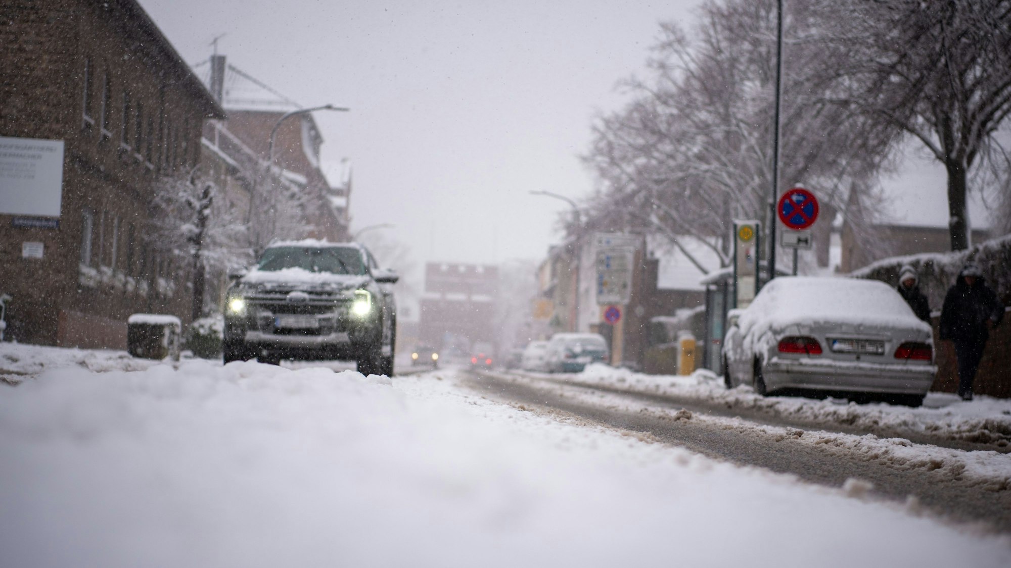 Ein Auto fährt durch Schneeflocken auf einer beschneiten Straße, im Hintergrund sind Häuser und weitere Autos zu erkennen.