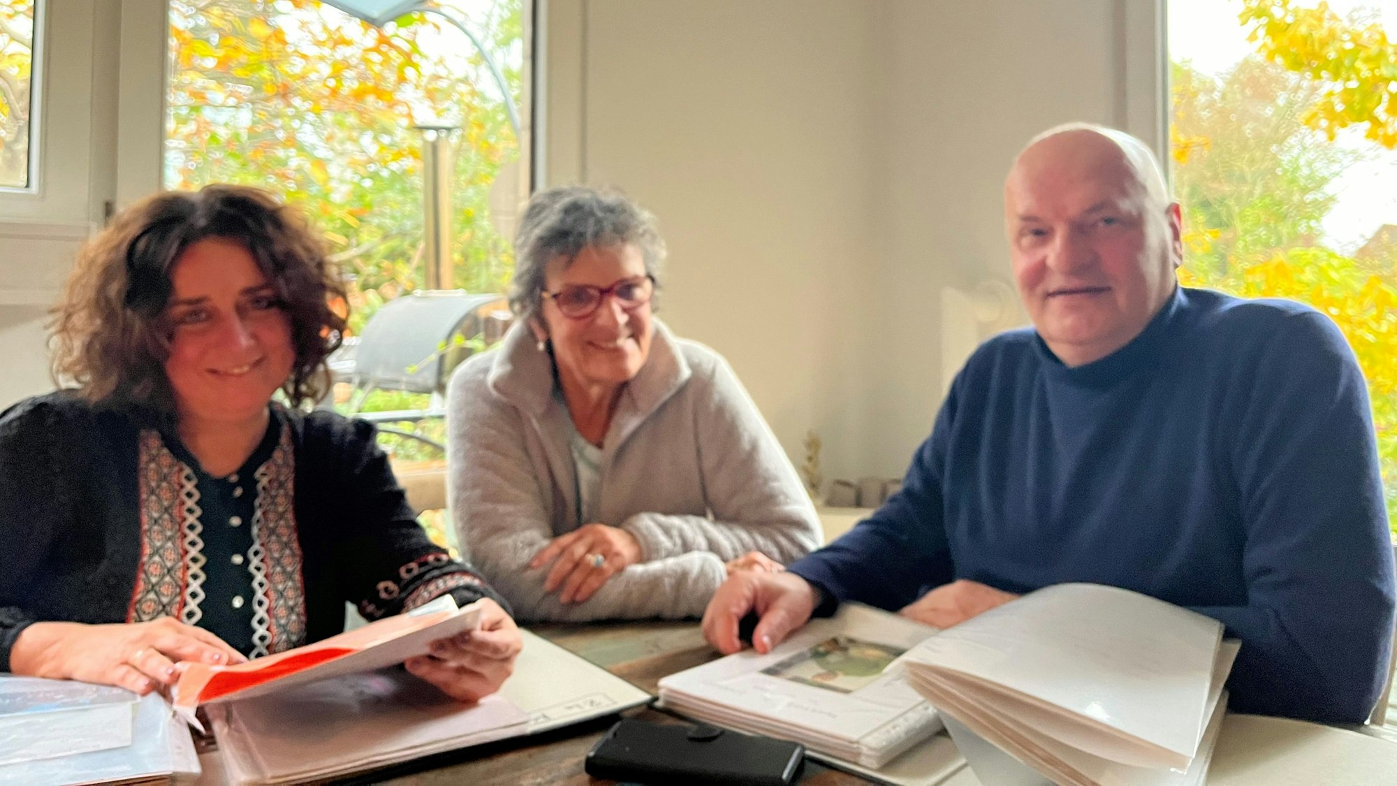 Die drei sitzen am Tisch, vor ihnen auf dem Tisch liegen Ordner.  und