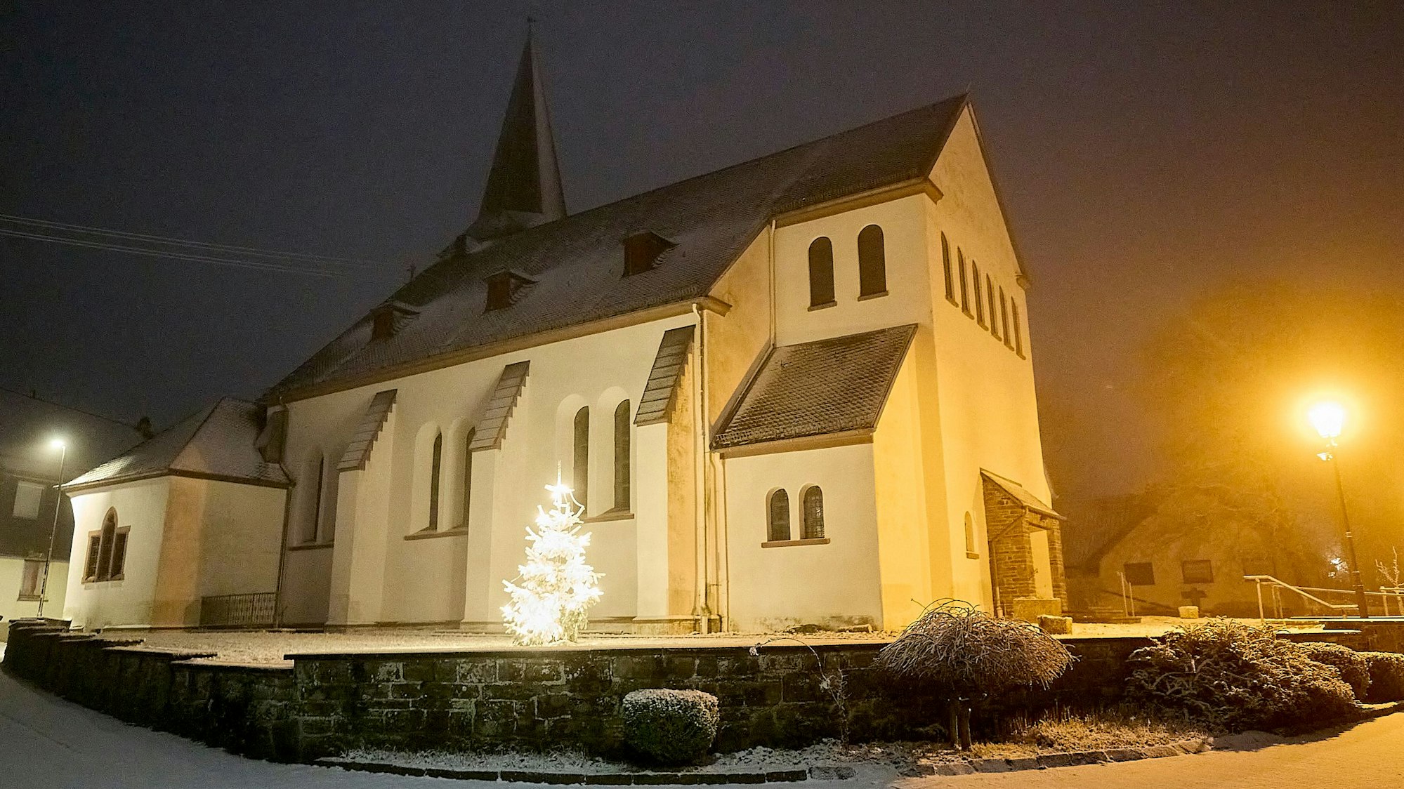 Die Aufnahme zeigt die von Straßenlaternen beleuchtete katholische Kirche in Hellenthal-Rescheid im Schnee. In der Dunkelheit leuchtet auch ein Weihnachtsbaum.