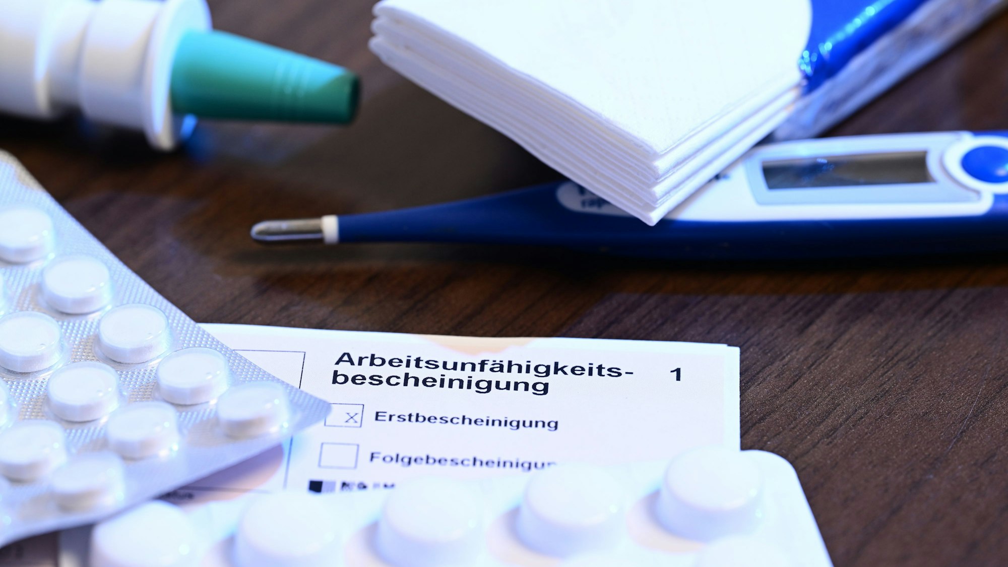 Ein Fieberthermometer, Medikamente und eine ärztliche Arbeitsunfähigkeitsbescheinigung (Krankmeldung) liegen auf einem Nachttisch (gestellte Szene).