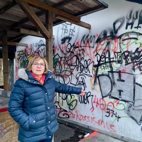 Eine Frau in einer dunkelblauen Steppjacke zeigt auf eine beschmierte Wand.