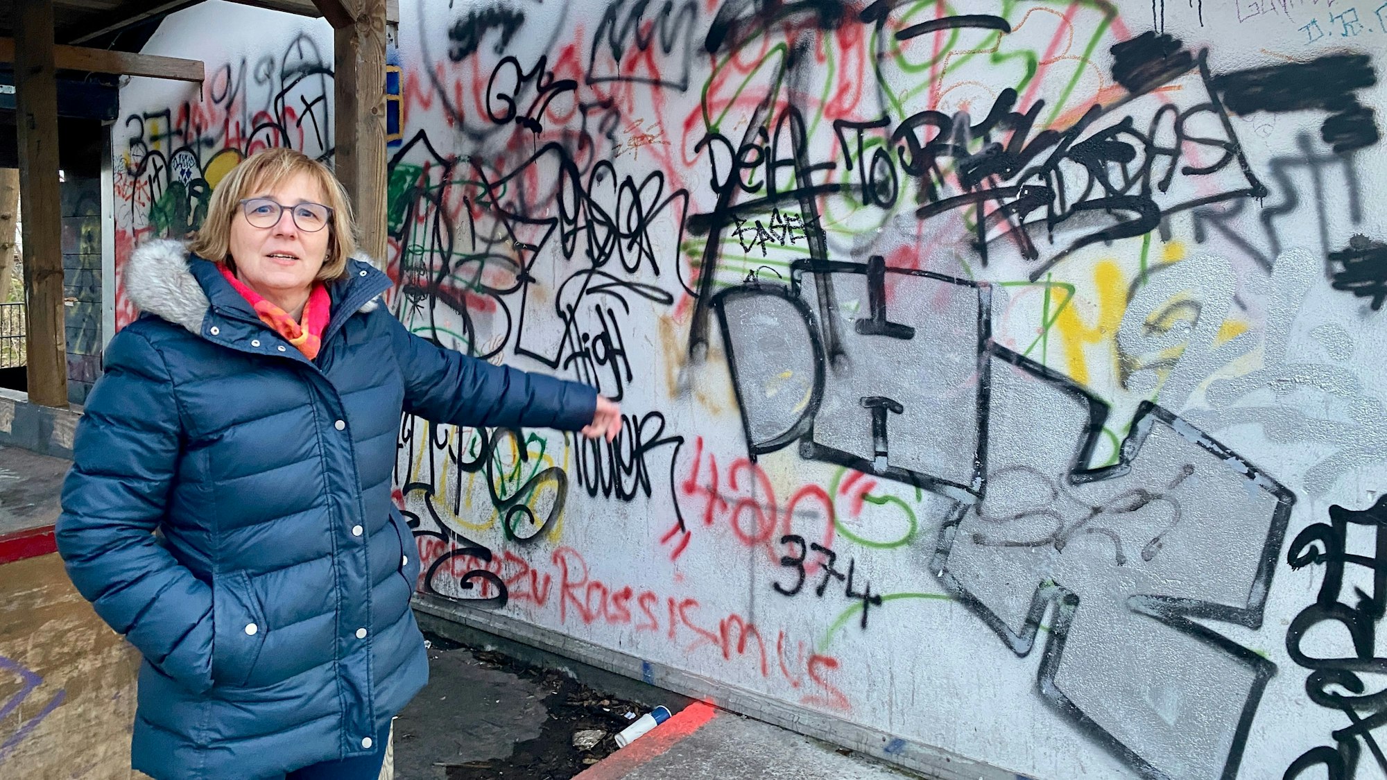 Eine Frau in einer dunkelblauen Steppjacke zeigt auf eine beschmierte Wand.