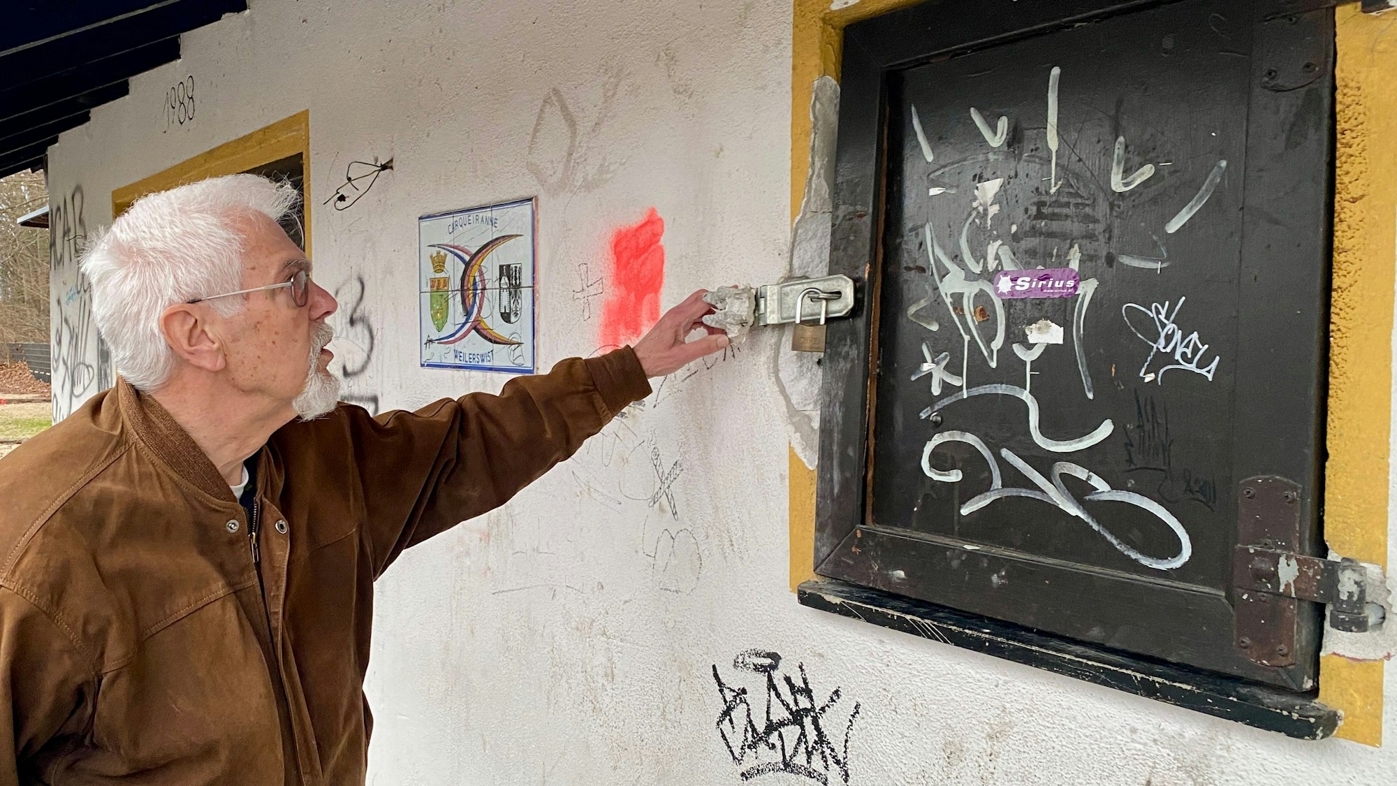 Ein Mann in brauner Lederjacke zeigt auf eine Verriegelung mit Vorhängeschloss an einem Fenster.
