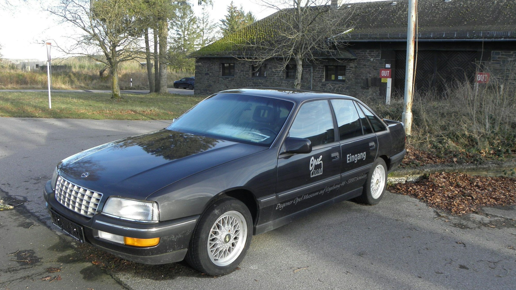 Ein schwarzes Auto, ein Opel Senator B, steht auf einem Parkplatz. Dahinter ist ein Haus.