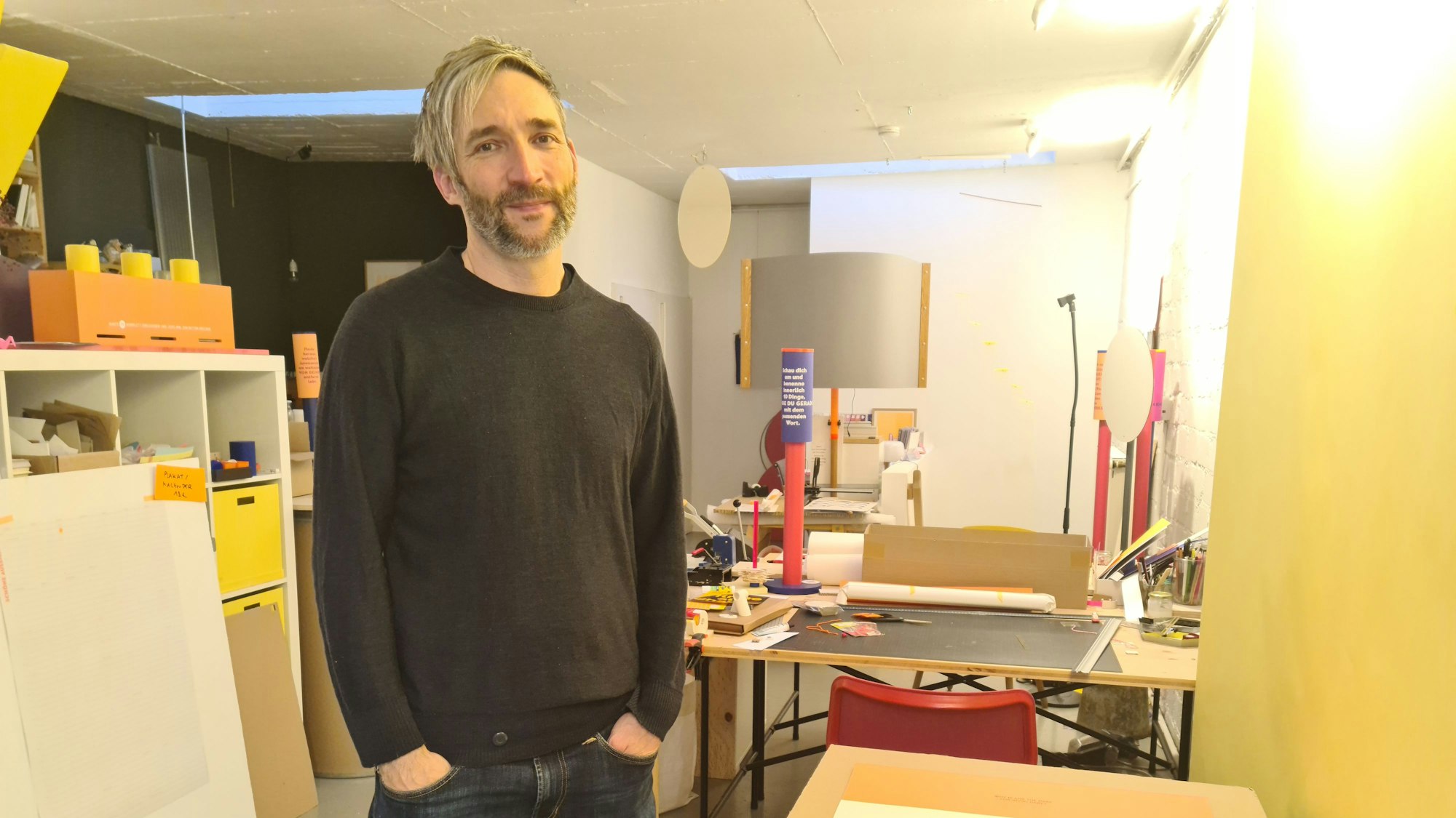 Ein Mann mit grauem Haar und Bart steht in einer Grafiker-Werkstatt neben einem Tisch.