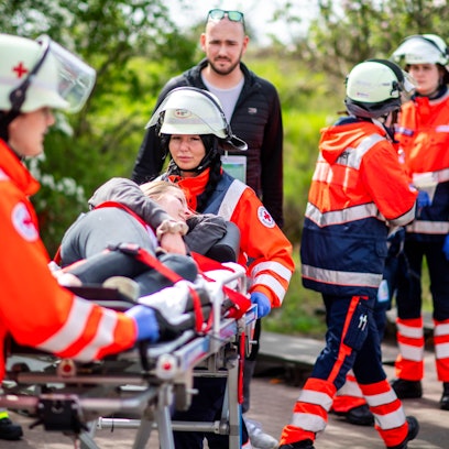 Eine Frau liegt auf einer Trage, die von zwei Rettungsdienstlern in roten Jacken geschoben wird. Im Hintergrund sind weiter Menschen in gelben und orangen Warnjacken zu sehen.