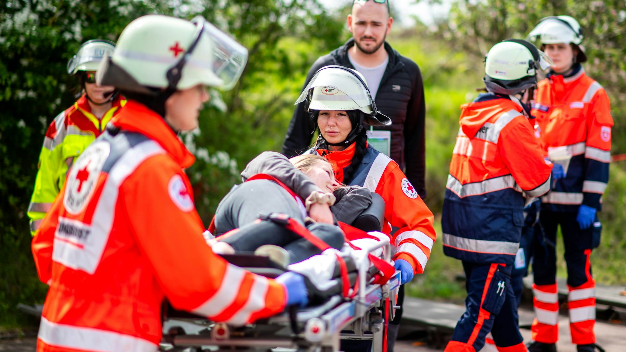 Eine Frau liegt auf einer Trage, die von zwei Rettungsdienstlern in roten Jacken geschoben wird. Im Hintergrund sind weiter Menschen in gelben und orangen Warnjacken zu sehen.