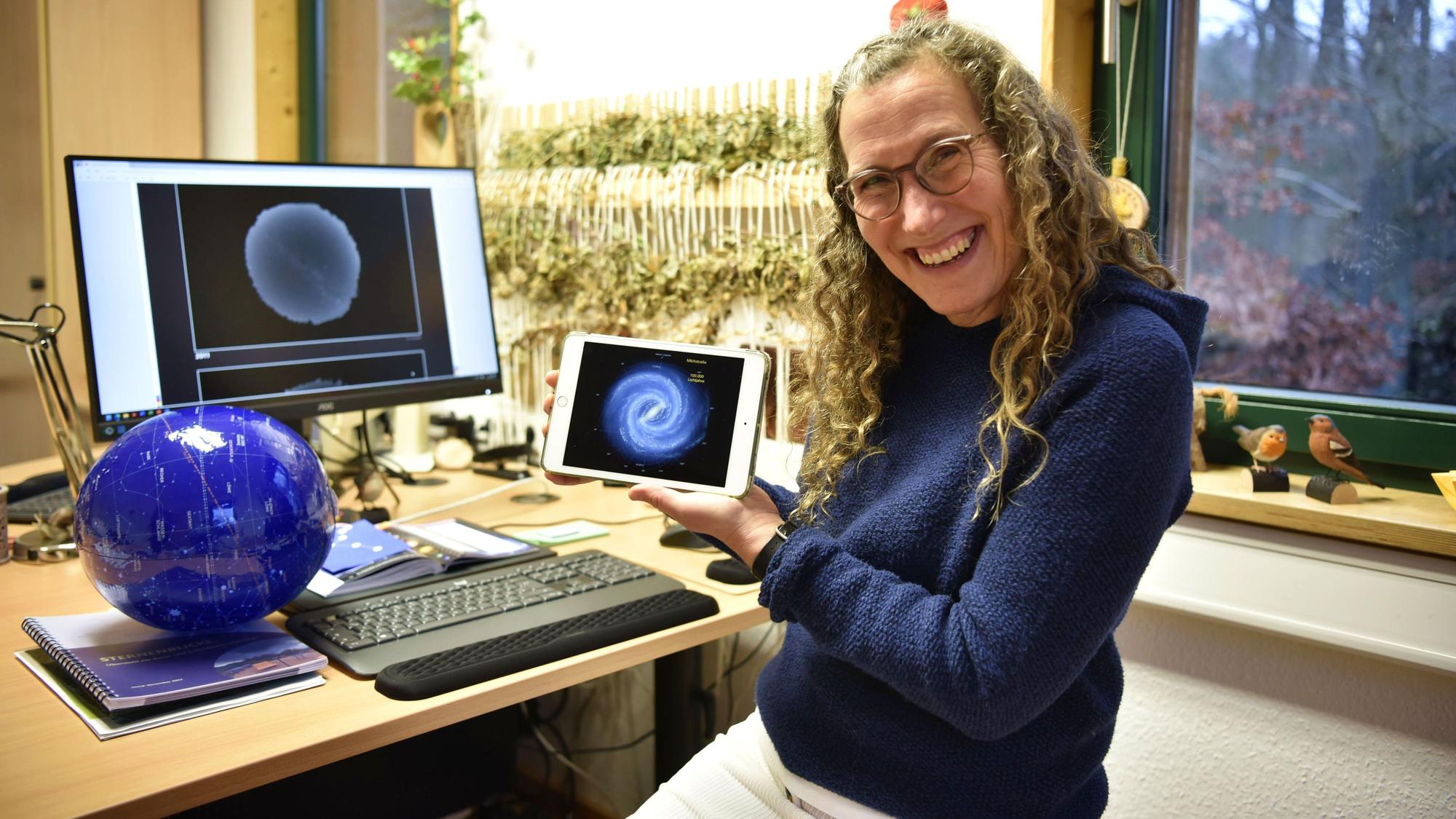 Michaela Wüller sitzt an ihrem Schreibtisch und zeigt auf einem kleinen Tablet die Milchstraße. Auf dem Schreibtisch liegen verschiedene Unterlagen zum Thema Sterne.