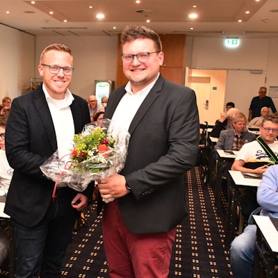 Das Foto zeigt zwei Männer mit einem Blumenstrauß vor besetzten Tischreihen.