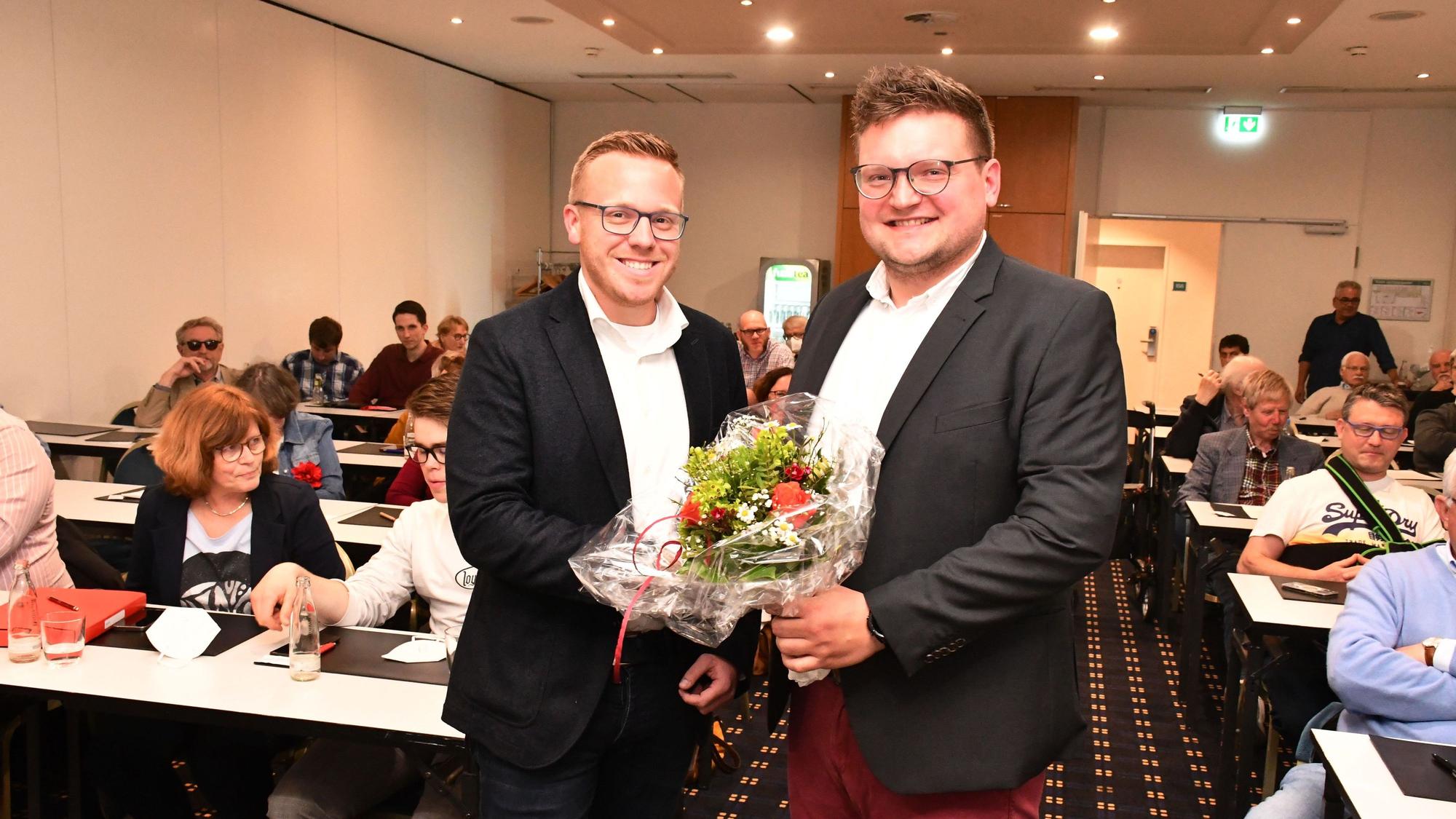 Das Foto zeigt zwei Männer mit einem Blumenstrauß vor besetzten Tischreihen.