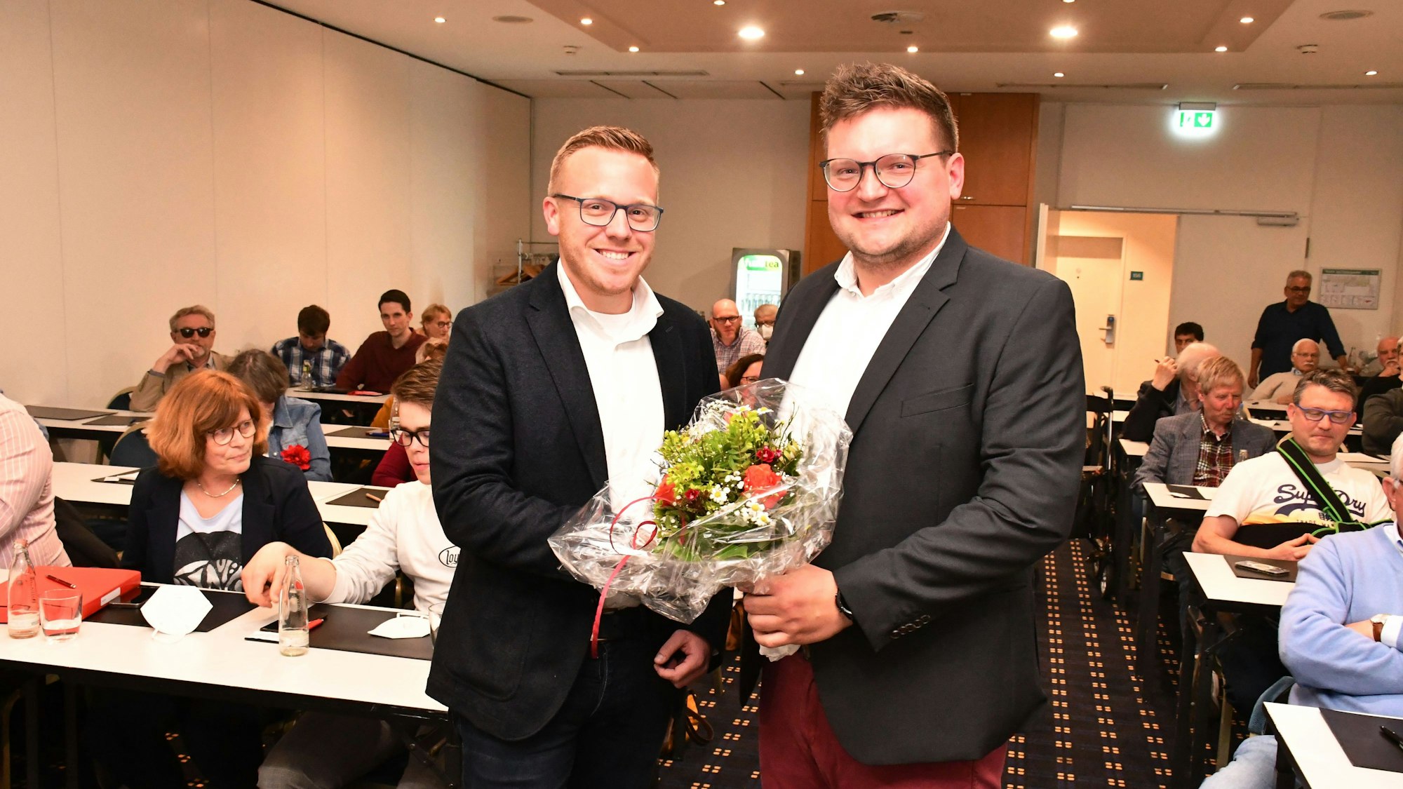 Das Foto zeigt zwei Männer mit einem Blumenstrauß vor besetzten Tischreihen.
