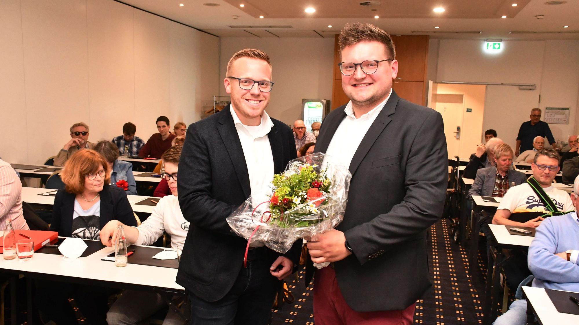 Das Foto zeigt zwei Männer mit einem Blumenstrauß vor besetzten Tischen in einem Saal.