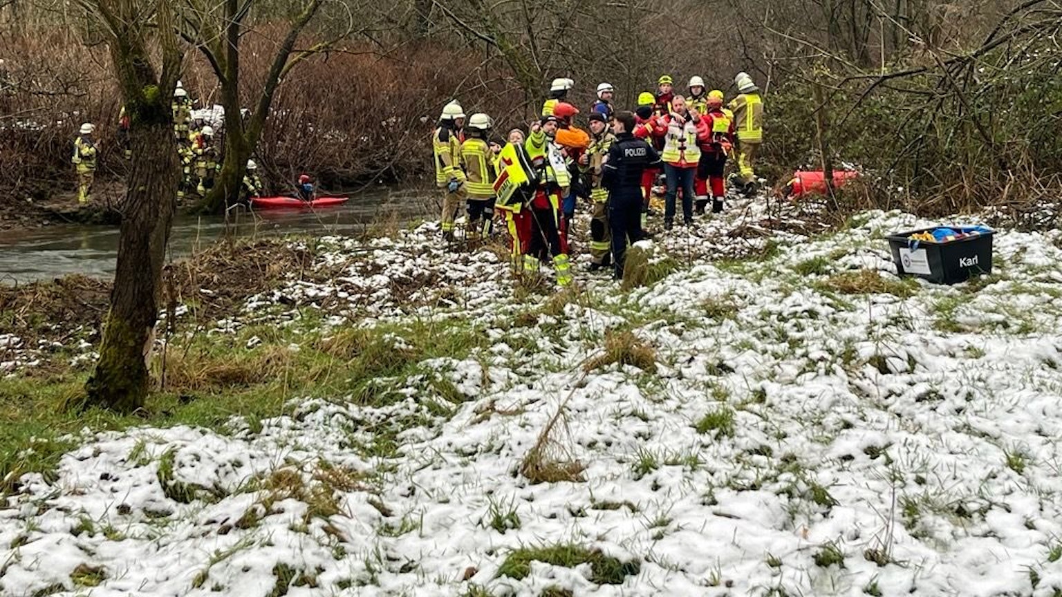 Rettungskräfte stehen an der Sülz auf einer schneebedeckten Wiese.