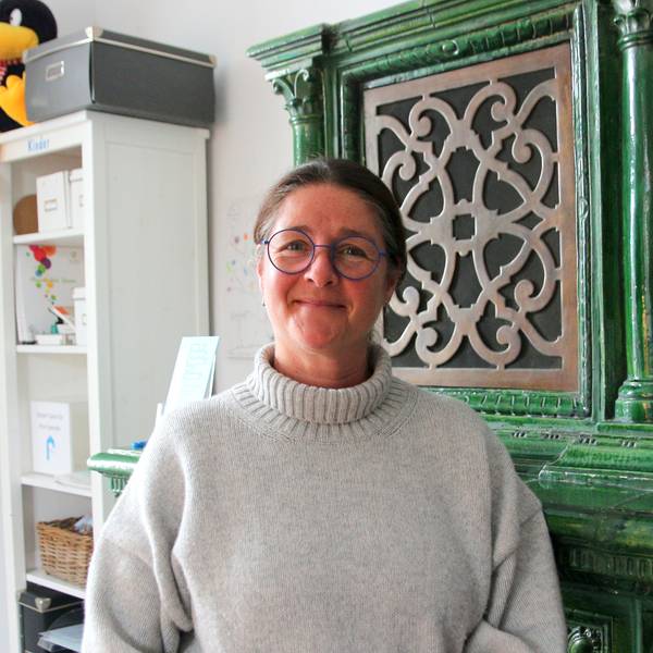 Auf dem Foto ist eine Frau zu sehen. Sie trägt einen hellen Rollkragenpullover und Brille. Sie steht vor einem antiken grünen Schrank.