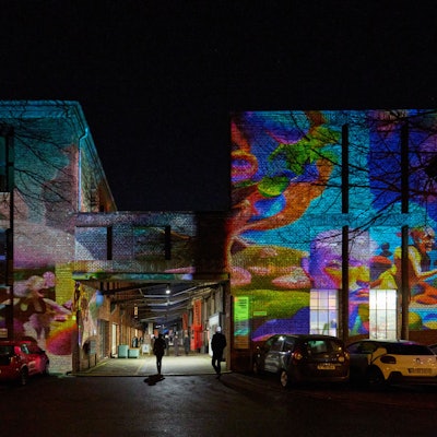 An der großen Backsteinwand des ehemaligen KHD-Gebäudes in Kalk küssen sich Menschen, bewegen sich Muster, wehen Äste im Wind.