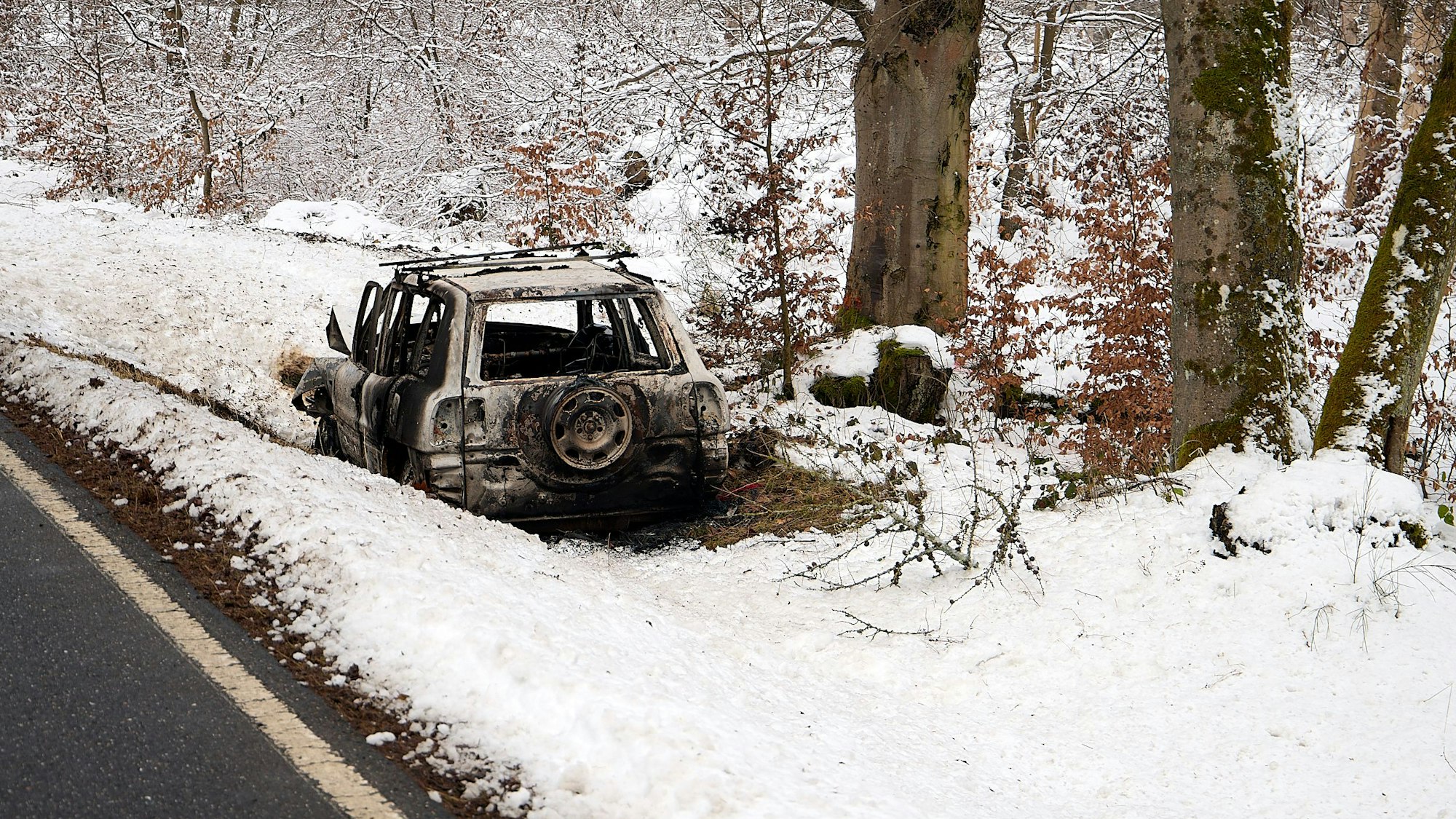 Ein ausgebranntes Fahrzeug steht im mit Schnee bedeckten Straßengraben neben der Kermeterstraße bei Schleiden-Wolfgarten.