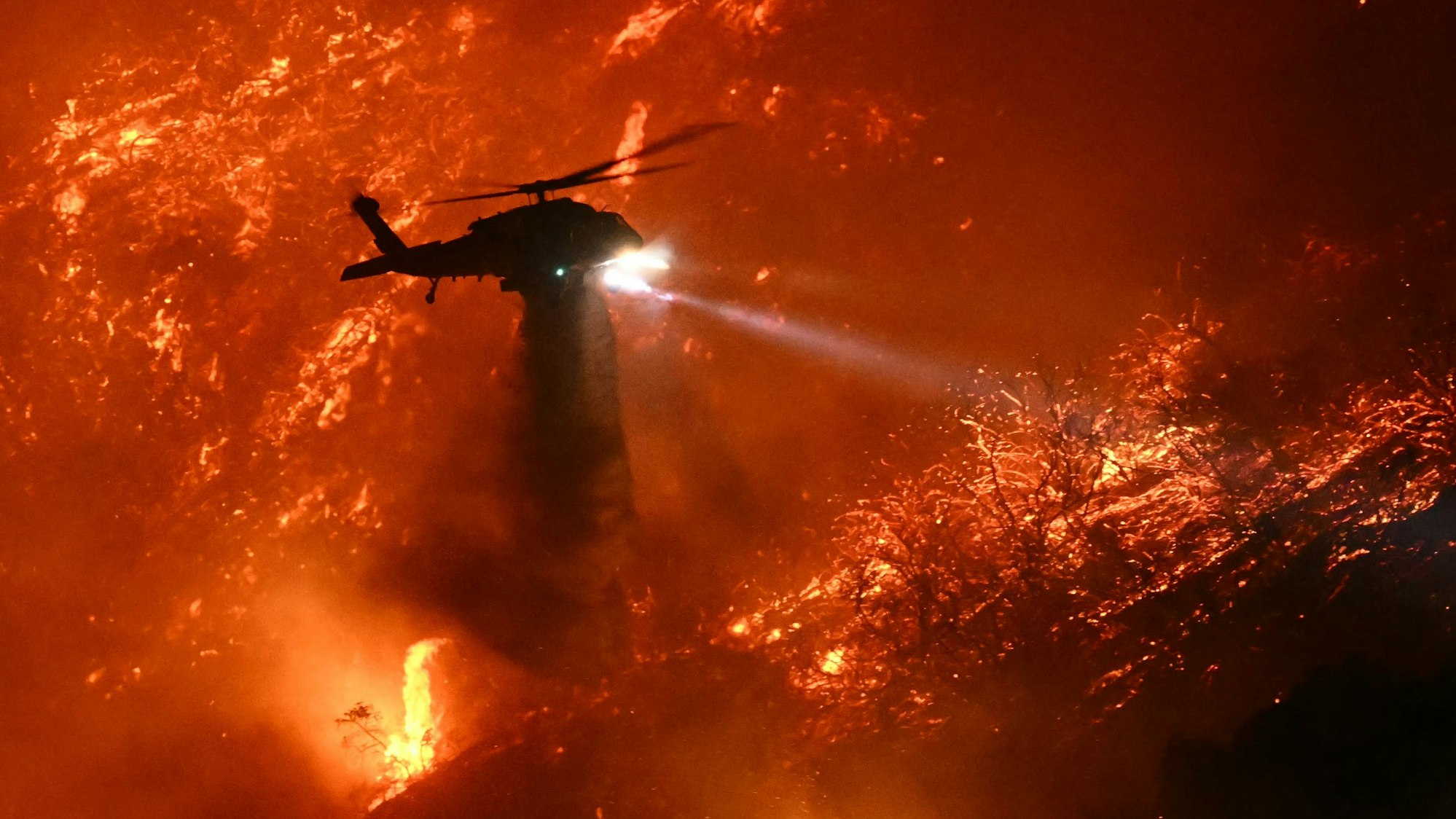 A fire fighting helicopter drops water as the Palisades fire grows near the Mandeville Canyon neighborhood and Encino, California, on January 11, 2025. The Palisades Fire, the largest of the Los Angeles fires, spread toward previously untouched neighborhoods January 11, forcing new evacuations and dimming hopes that the disaster was coming under control. Across the city, at least 11 people have died as multiple fires have ripped through residential areas since January 7, razing thousands of homes in destruction that US President Joe Biden likened to a "war scene." (Photo by Patrick T. Fallon / AFP)