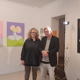 Eine Frau und ein Mann stehen zwischen Kunstwerken, die links und rechts von ihnen an der Wand hängen.