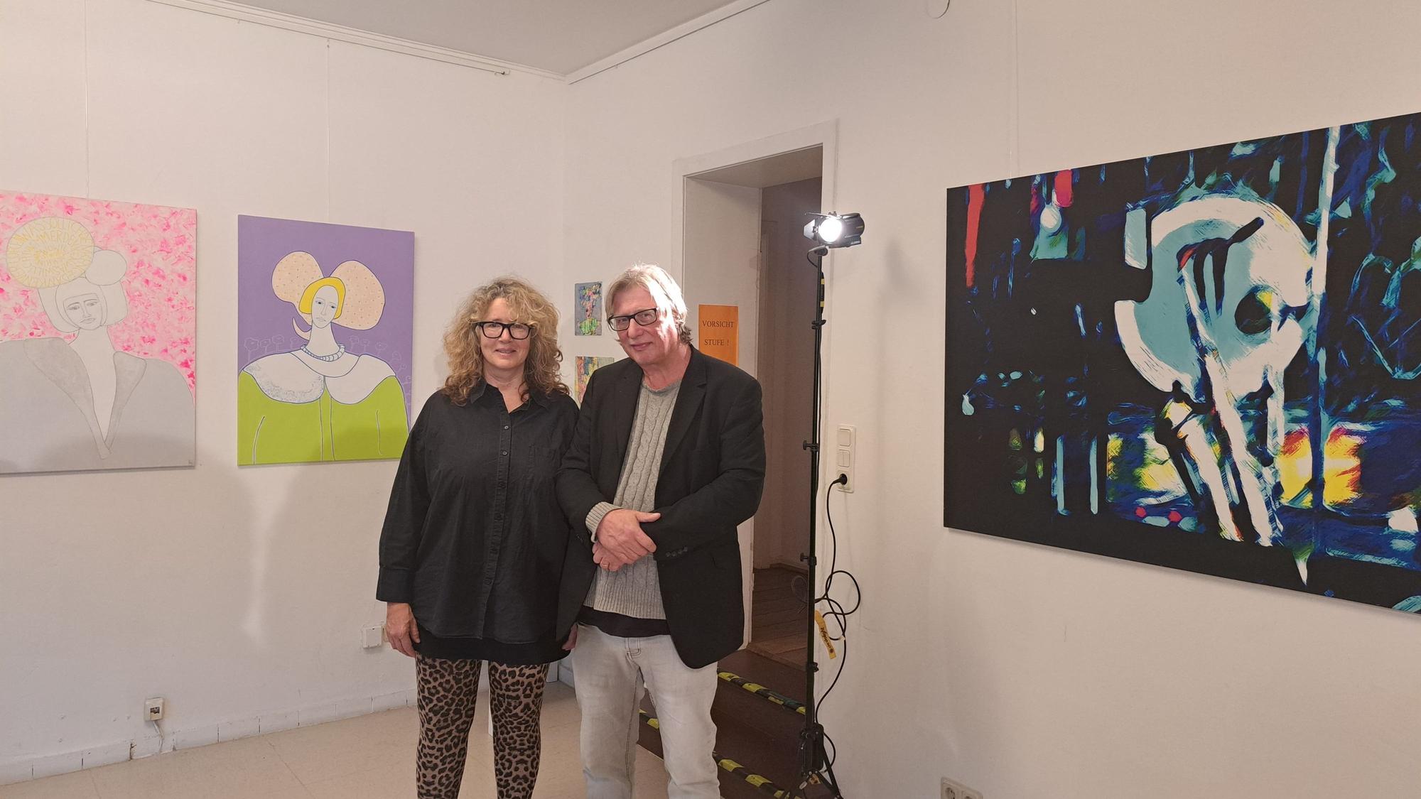 Eine Frau und ein Mann stehen zwischen Kunstwerken, die links und rechts von ihnen an der Wand hängen.