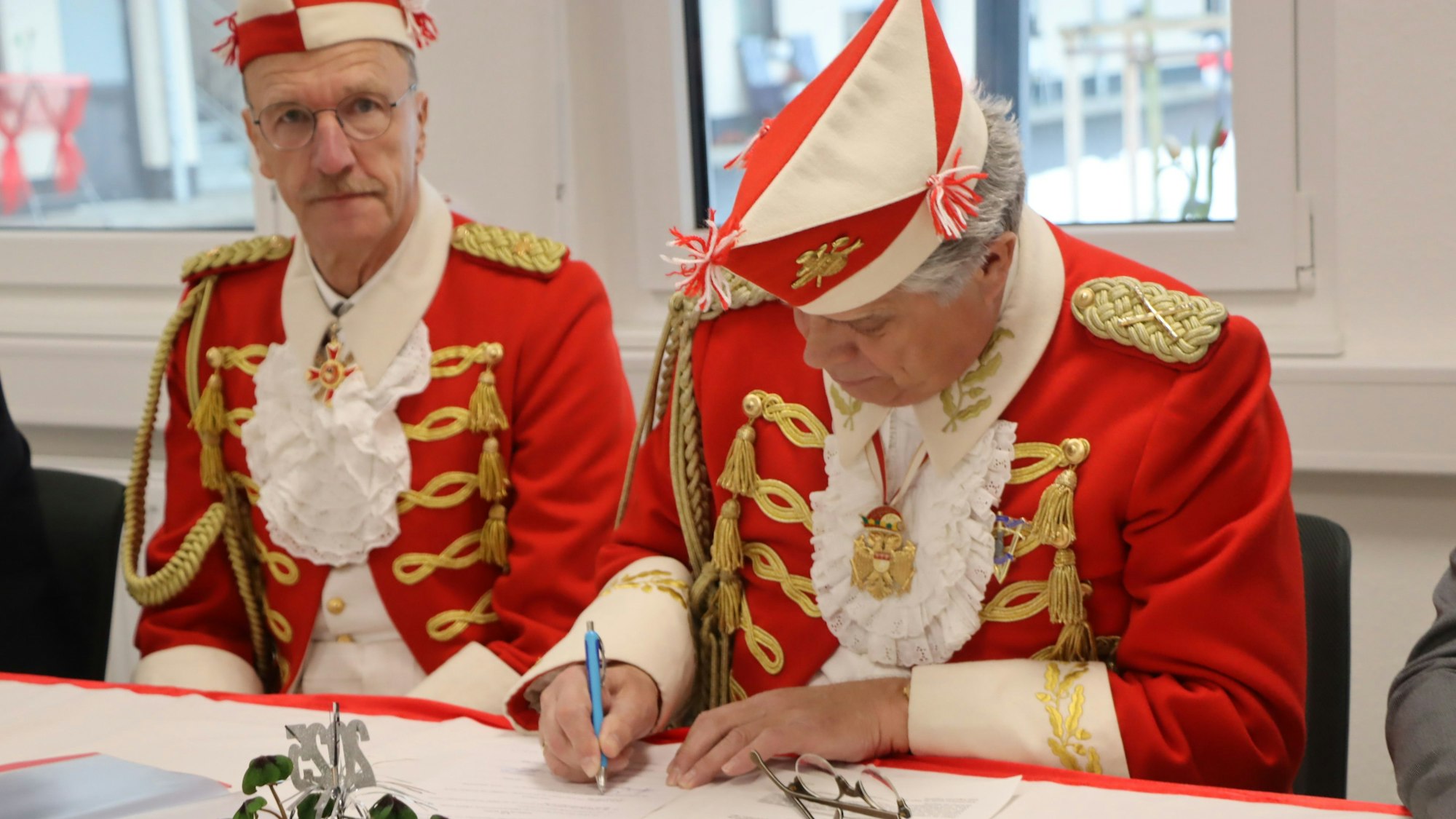 Zwei Männer in rot-weißen Uniformen sitzen an einem Tisch, einer unterschreibt ein Dokument.