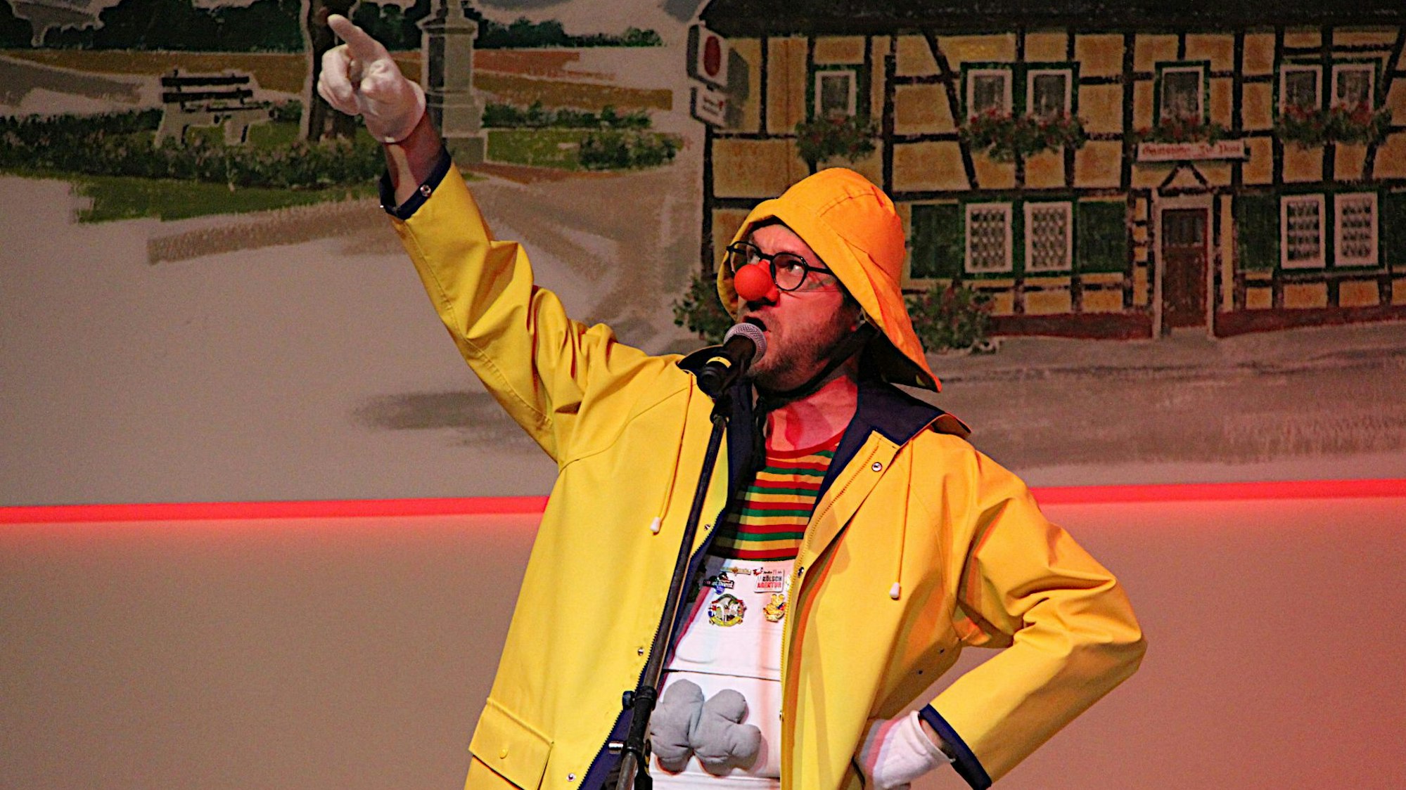 Björn Wassong steht in gelbem Regenmantel und Hut sowie einer roten Pappnase auf der Bühne in Zülpich-Dürscheven und hält eine Büttenrede.