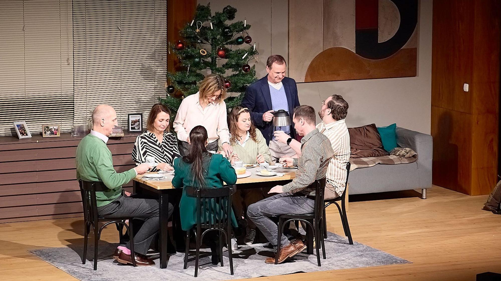 Auf der Theaterbühne sieht man acht Männer und Frauen unterschiedlichen Alters an einer Kaffeetafel. Im Hintergrund steht ein geschmückter Weihnachtsbaum.