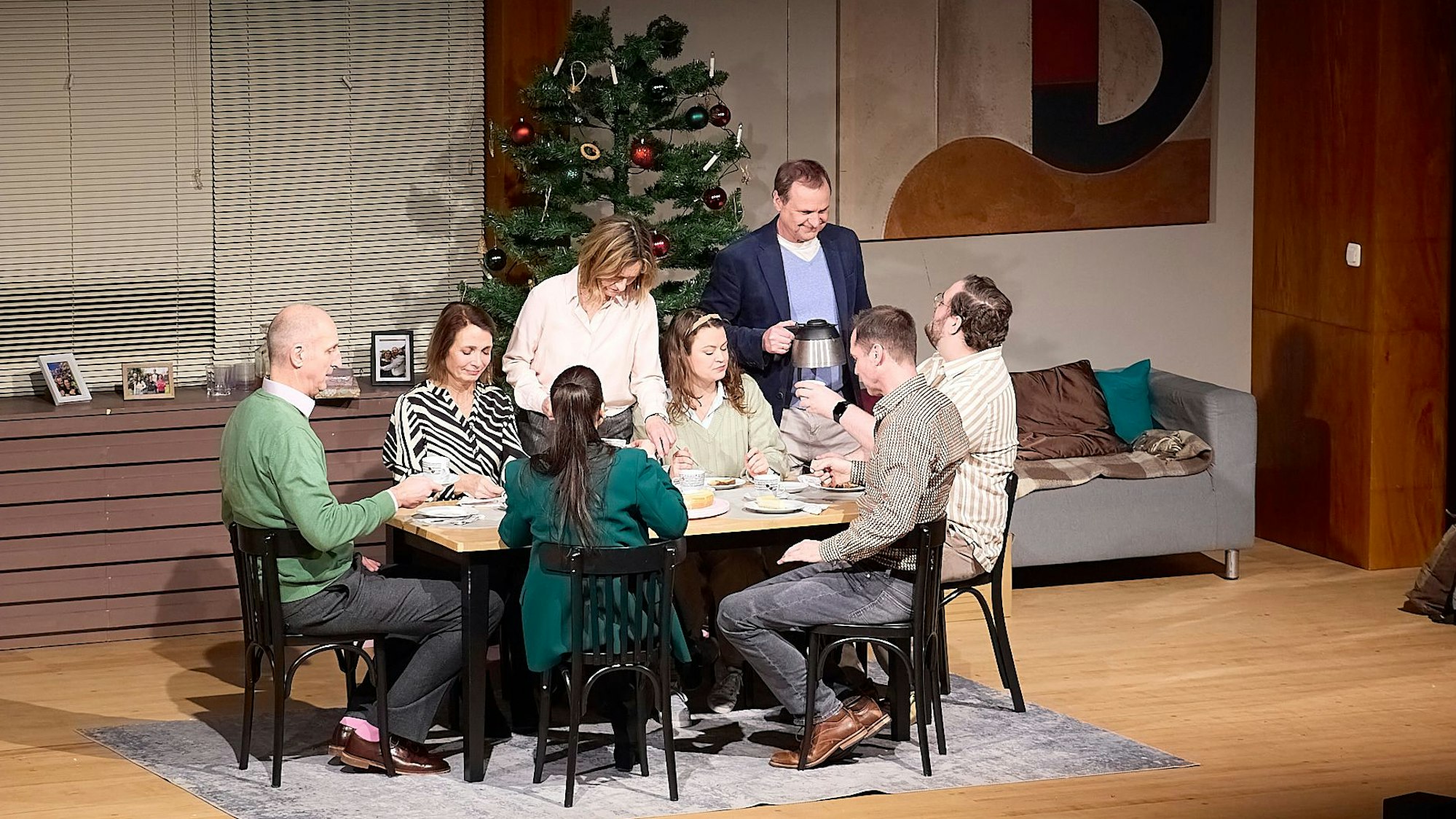 Auf der Theaterbühne sieht man acht Männer und Frauen unterschiedlichen Alters an einer Kaffeetafel. Im Hintergrund steht ein geschmückter Weihnachtsbaum.