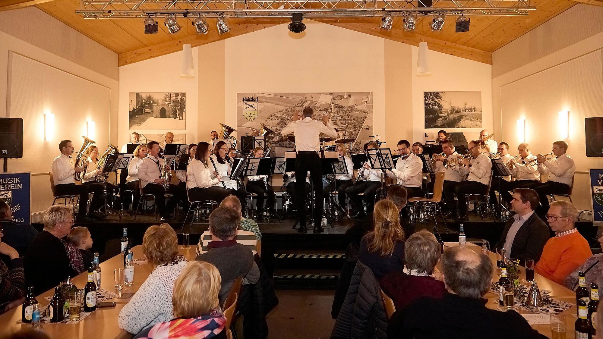 Im Dorfsaal Floisdorf stehen die Musiker des Musikvereins Cäcilia Floisdorf aufv der Bühne. Das Publikum sitzt an den Tischen und lauscht.