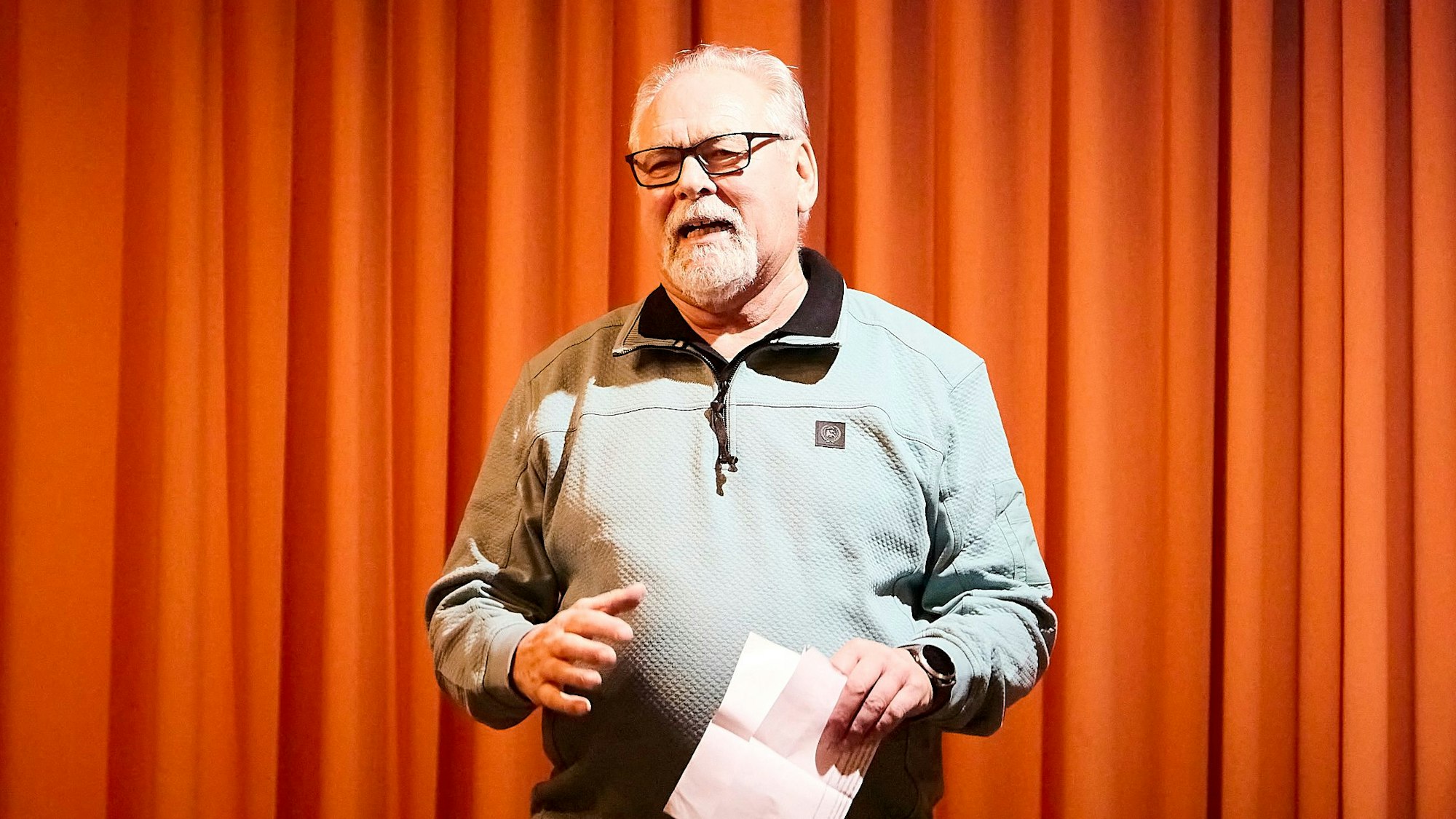 Ortsvorsteher Robert Ohlerth steht vor dem orangefarbenen Vorhang der Bühne.