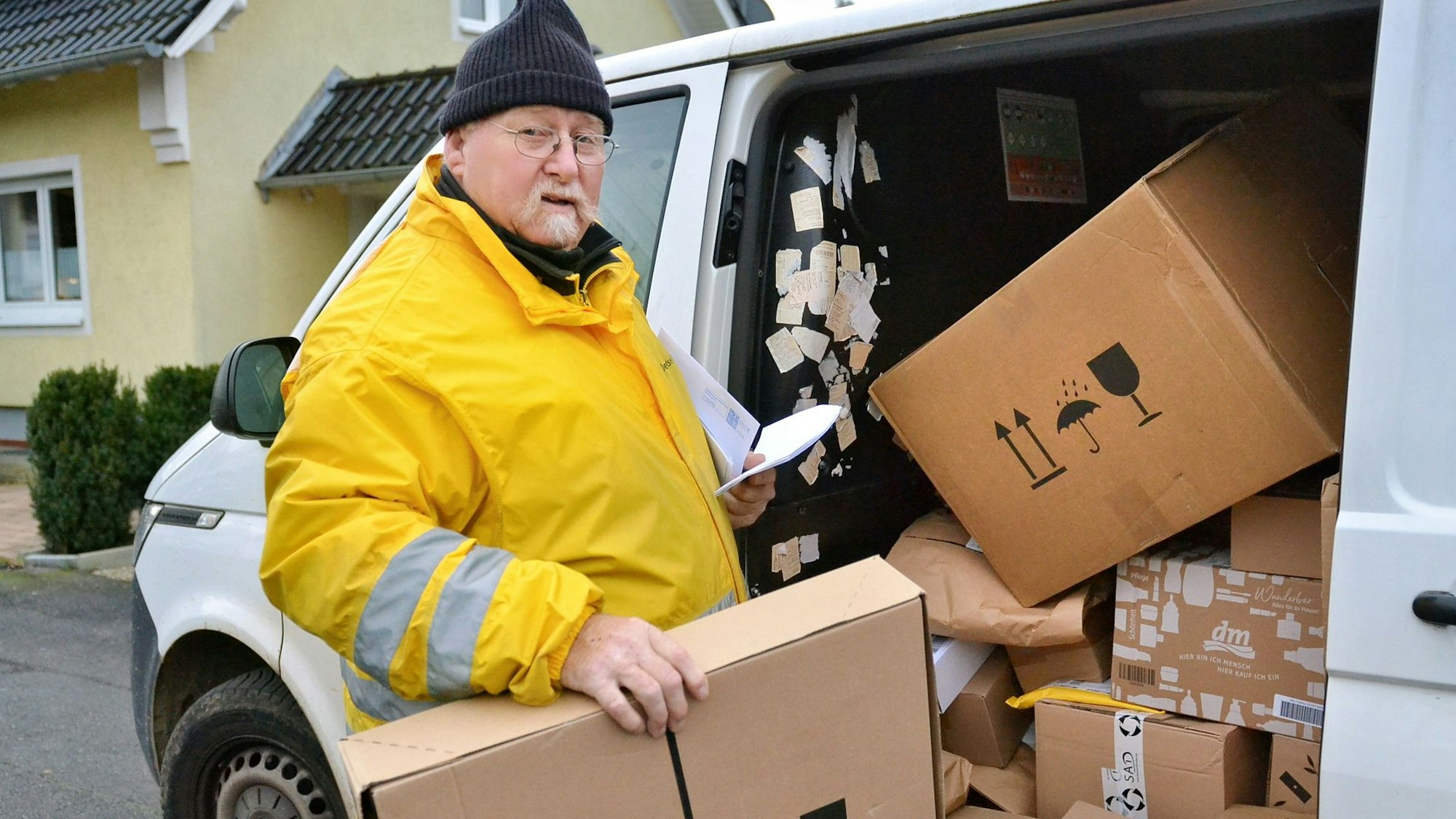 Zusteller Ralf Junkers trägt eine gelbe Jacke und steht an seinem geöffneten Lieferfahrzeug. Darin stapeln sich etliche Pakete.