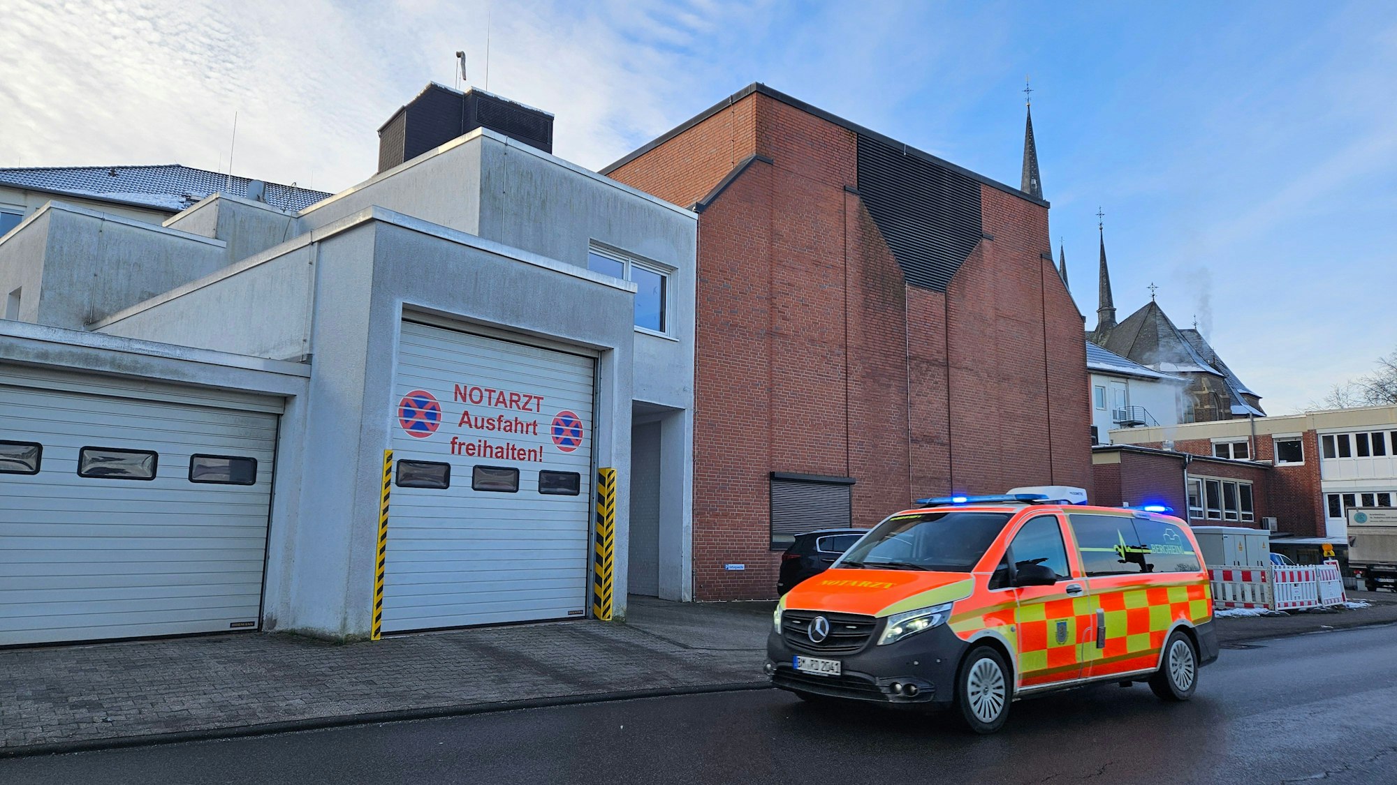 Zu sehen ist das Krankenhaus in Bedburg, im Vordergrund das Fahrzeug des Notarztes im Einsatz mit Blaulicht.