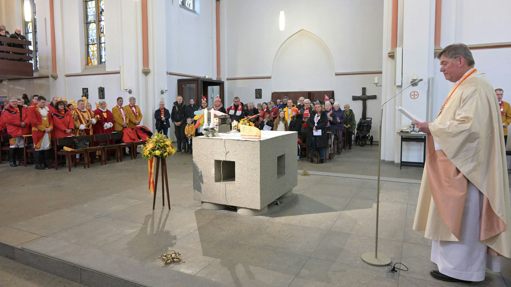 Ein katholischer Pfarrer steht am Mikrofon in einer Kirche. Man sieht einen modernen Altar-Quader, in den Bänken stehend zahlreiche Gottesdienstbesucher.