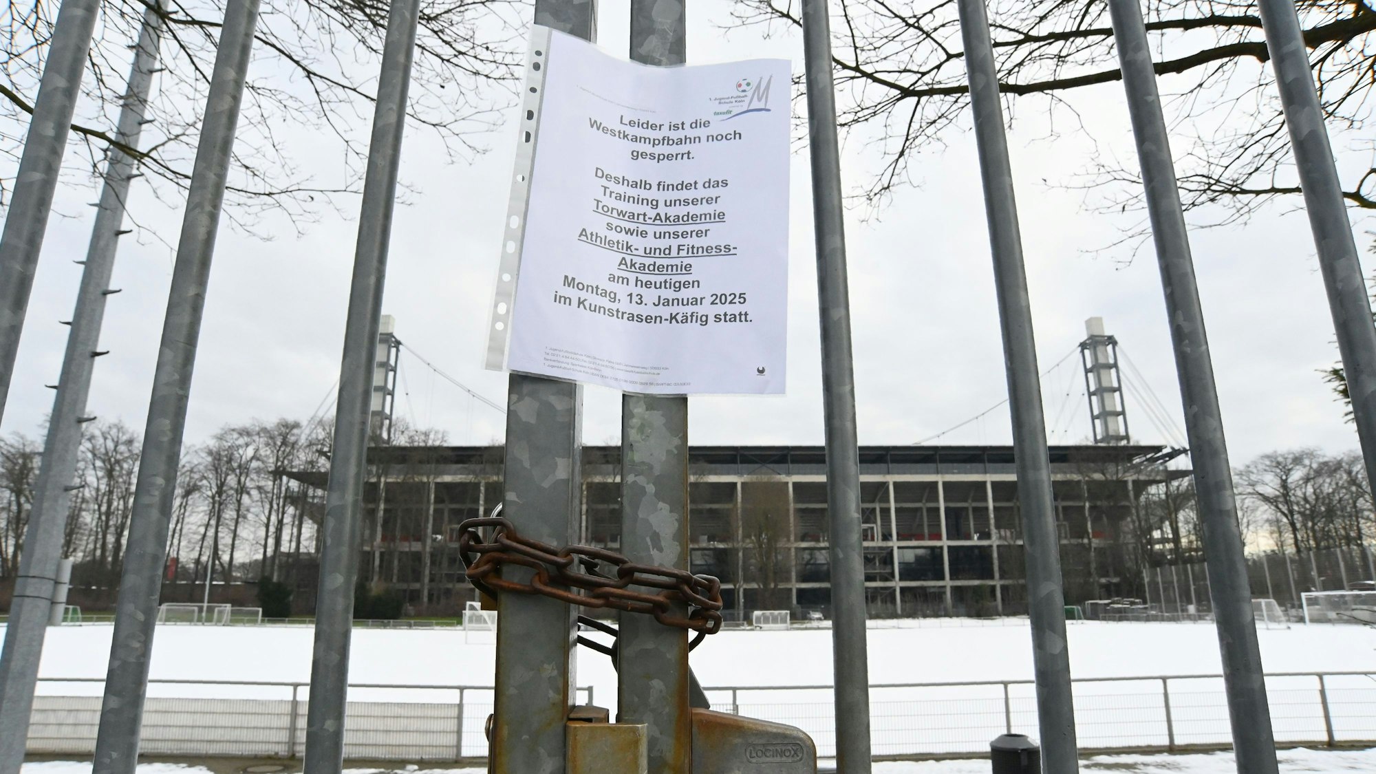 Die verschneite Kunstrasenfläche der Westkampfbahn vor dem Rhein-Energie-Stadion liegt hinter einem Gitter. Ein Schild deutet auf die Sperrung der Fläche hin.