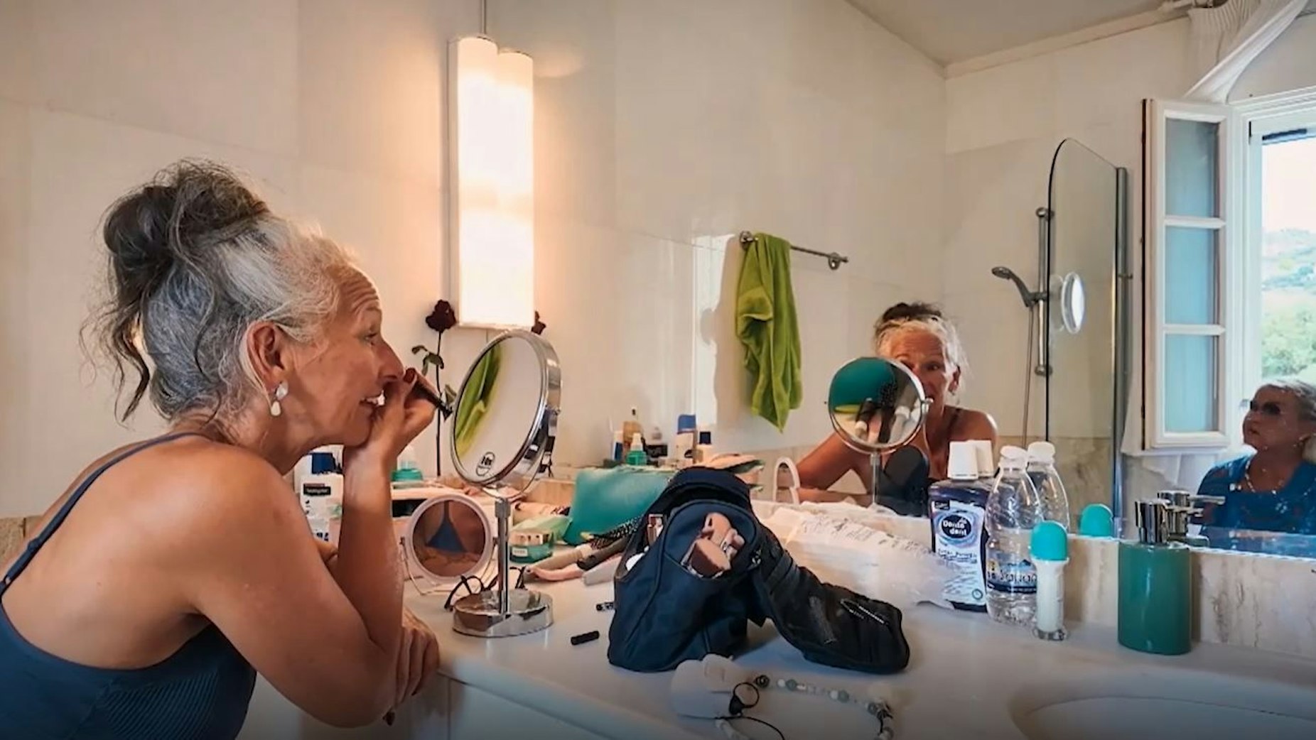 Vorbereitung auf das Einzeldate mit Franz: Sabine schminkt sich vor einem Spiegel. Eine Frau rechts im Bild schaut ihr durch eine Sonnenbrille dabei zu.