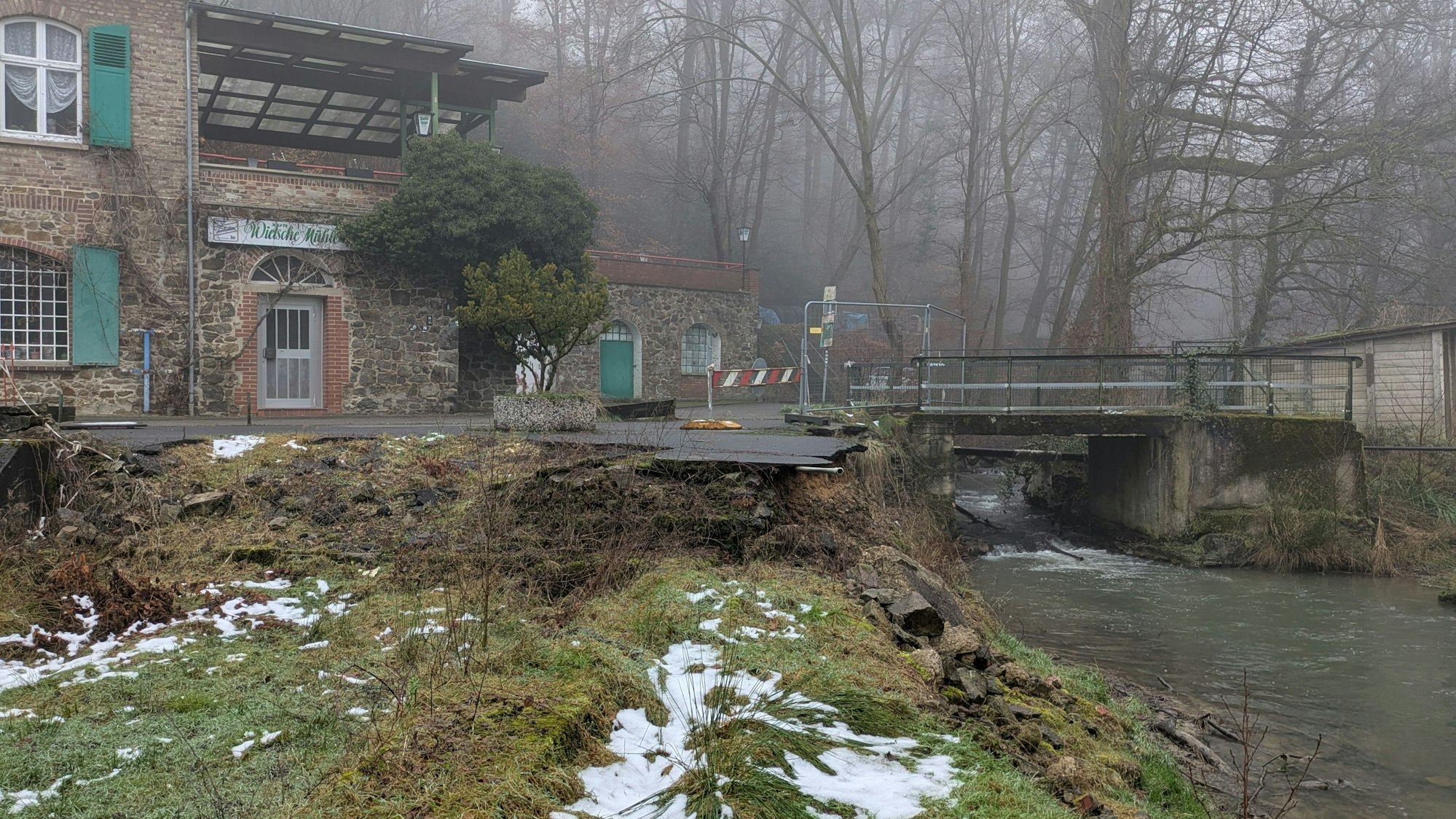 Eine alte, nicht mehr benutzbare Brücke über einen Bach neben einem Gebäude.