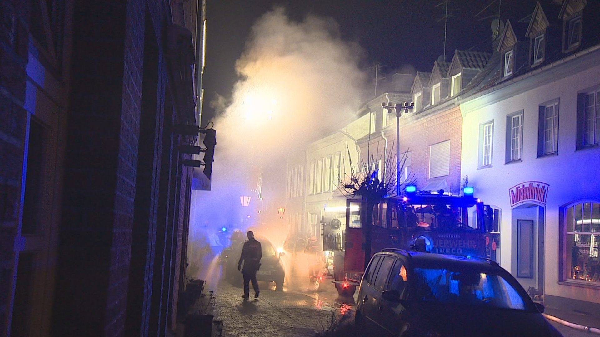 Einsatzkräfte bekämpfen einen Wohnungsbrand in Kalkar. Eine kleine Straße ist in der Nacht mit Rauch gefüllt, davor ein Feuerwehrauto mit Blaulicht.