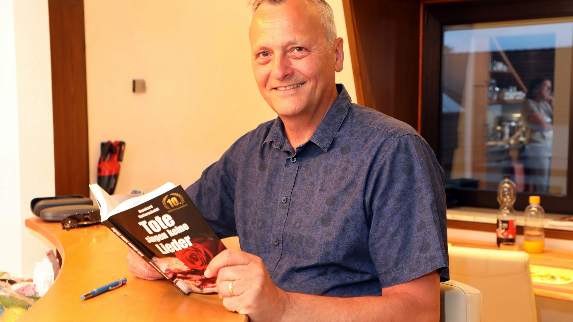 Autor Bernhard Hatterscheid sitzt mit einem Roman an der Küchenbar seines Hauses in Oberodenthal.