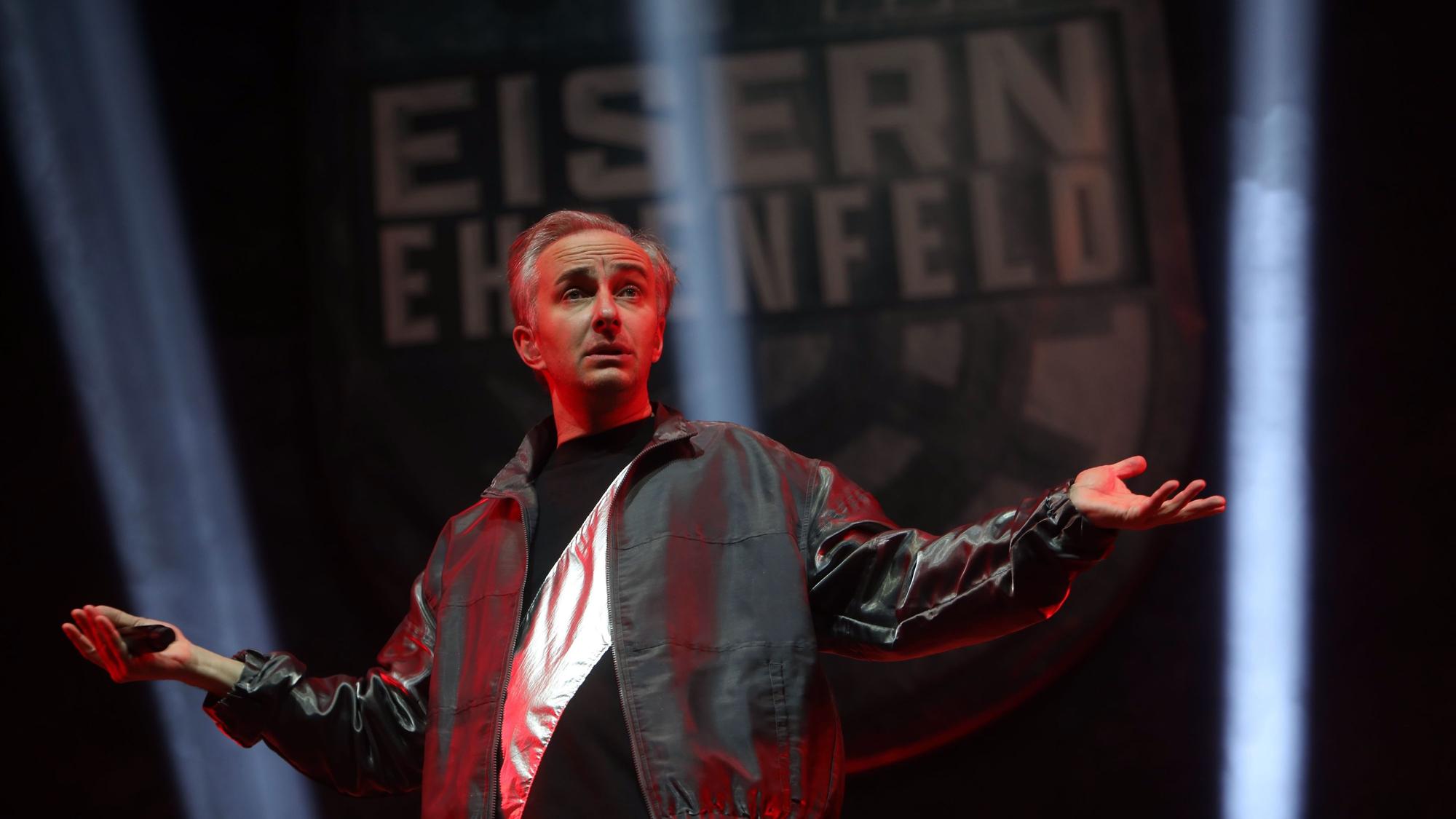 14.01.2025, Köln: Jan Böhmermann & Das Rundfunk Tanzorchester Ehrenfeld in der Lanxess Arena. Böhmermann hat die Arme ausgebreitet, trägt eine dunkle Silberjacke.