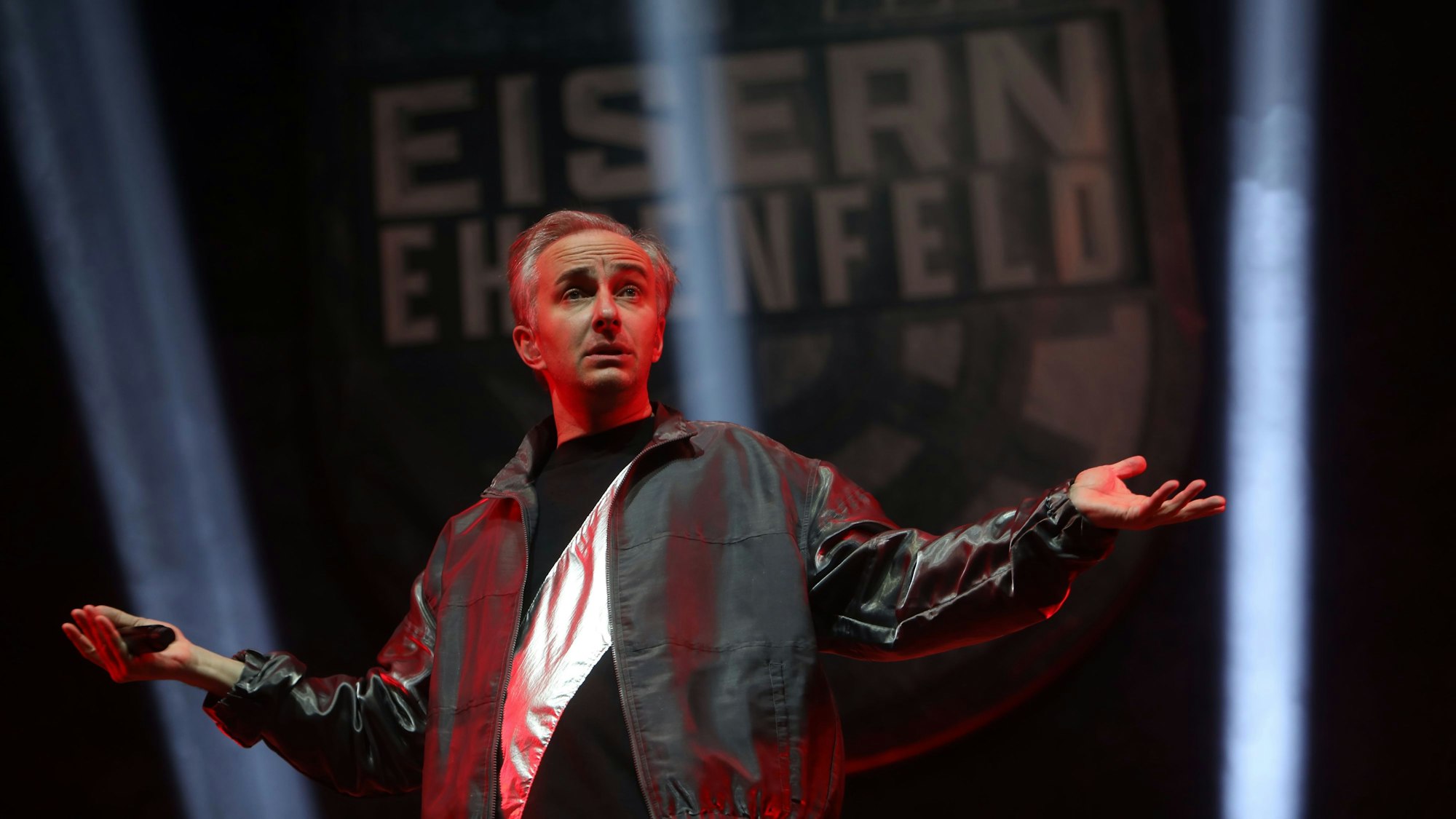 14.01.2025, Köln: Jan Böhmermann & Das Rundfunk Tanzorchester Ehrenfeld in der Lanxess Arena. Böhmermann hat die Arme ausgebreitet, trägt eine dunkle Silberjacke.