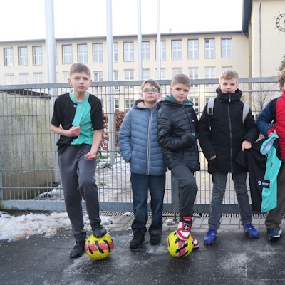 Sechs Jungen stehen mit drei Fußbällen vor einem verschlossenen Tor. Im Hintergrund ist eine Schule zu erkennen.