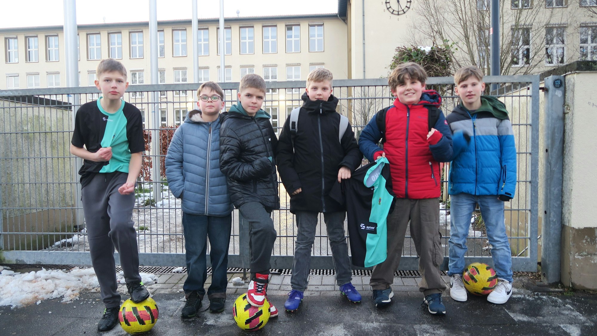Sechs Jungen stehen mit drei Fußbällen vor einem verschlossenen Tor. Im Hintergrund ist eine Schule zu erkennen.