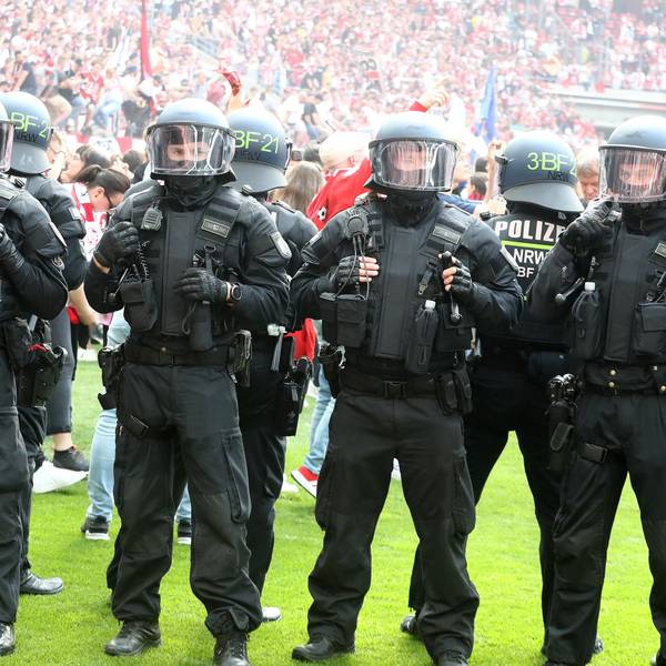 Polizisten stehen in Reihe auf dem Rasen eines Fußballstadions und hindern Fans daran, das Spielfeld einzunehmen. Die Polizisten tragen Schutzkleidung sowie Helme.