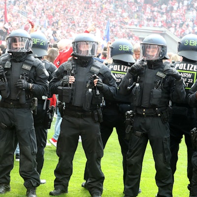 Polizisten stehen in Reihe auf dem Rasen eines Fußballstadions und hindern Fans daran, das Spielfeld einzunehmen. Die Polizisten tragen Schutzkleidung sowie Helme.