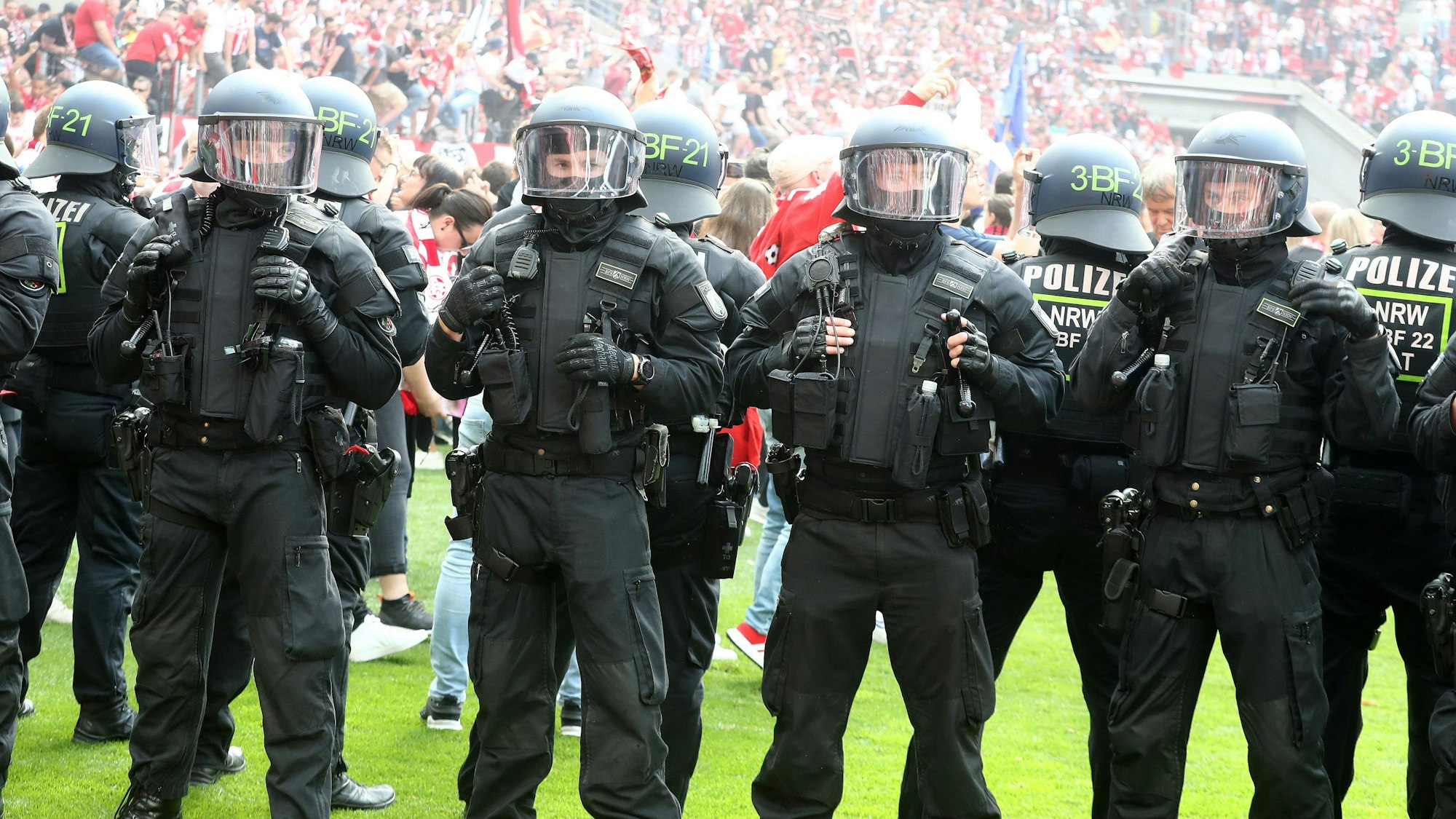 Polizisten stehen in Reihe auf dem Rasen eines Fußballstadions und hindern Fans daran, das Spielfeld einzunehmen. Die Polizisten tragen Schutzkleidung sowie Helme.