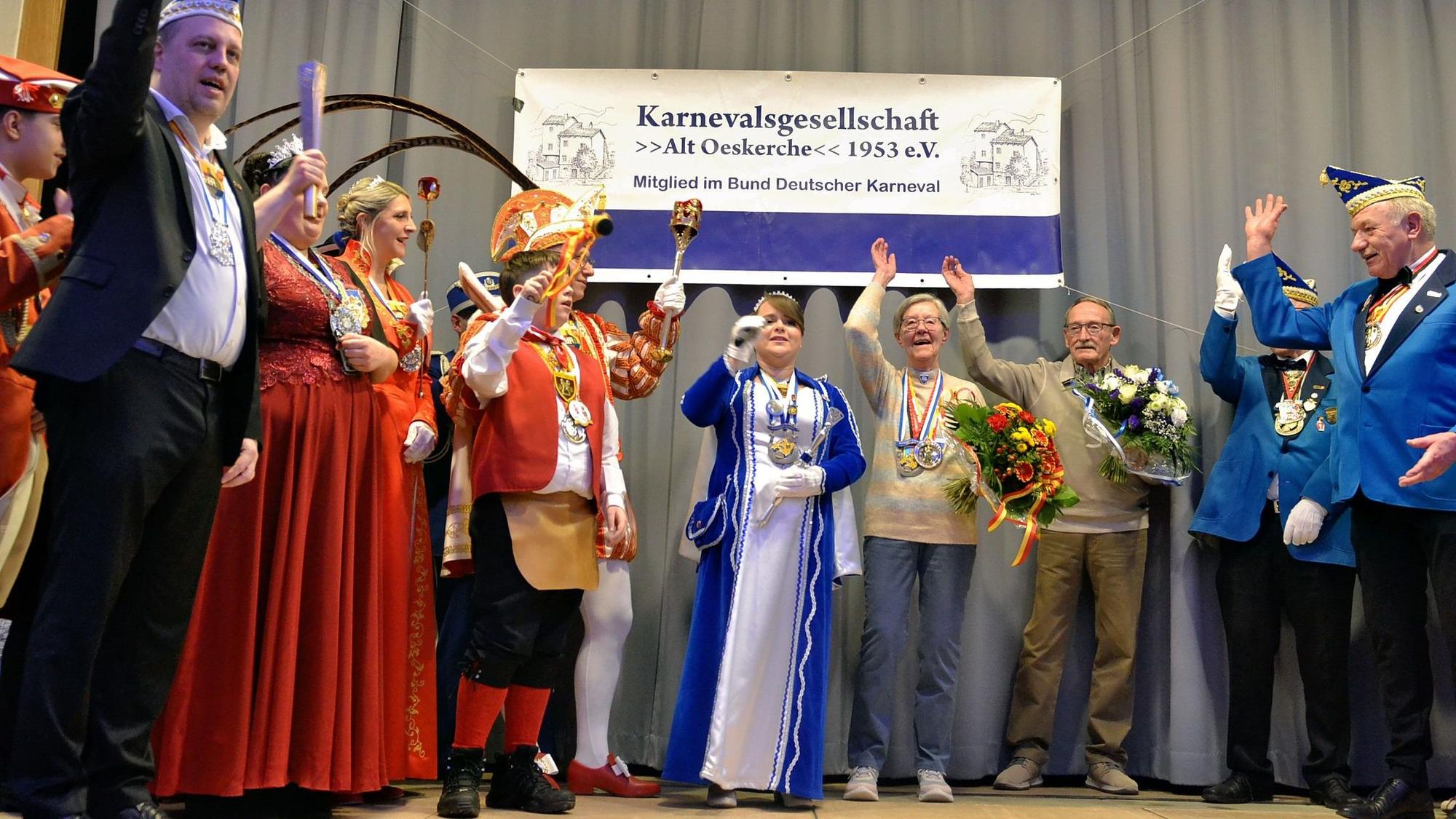 Auf einer Bühne stehen Karnevalisten unterschiedlicher Vereine und heben ihre Arme zum Alaaf-Ruf. In ihrer Mitte steht die Ordensträgerin mit ihrem Lebensgefährten. Beide halten Blumensträuße in den Händen.
