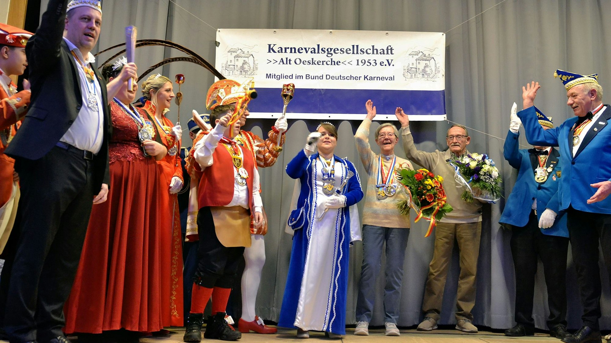 Auf einer Bühne stehen Karnevalisten unterschiedlicher Vereine und heben ihre Arme zum Alaaf-Ruf. In ihrer Mitte steht die Ordensträgerin mit ihrem Lebensgefährten. Beide halten Blumensträuße in den Händen.